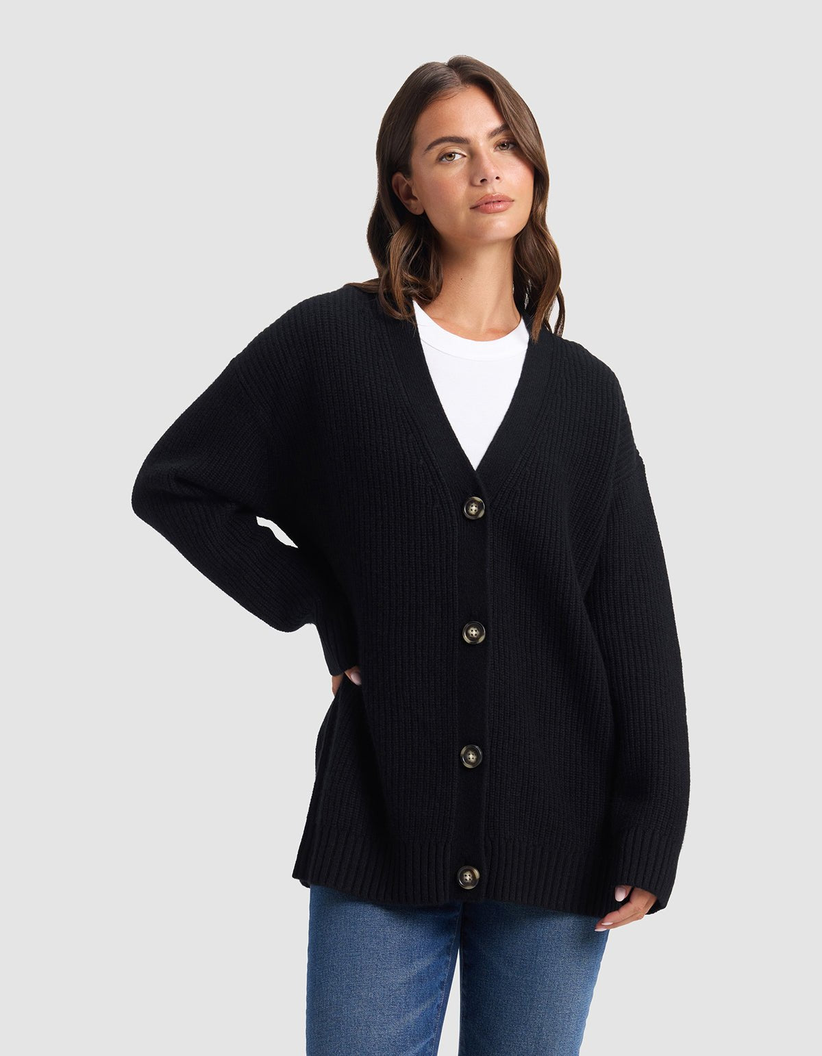A woman stands against a light gray background, looking to the side with one hand on her hip, wearing Cozy Earth's Oversized Sunday Cashmere Cardigan in black over a white shirt and blue jeans.