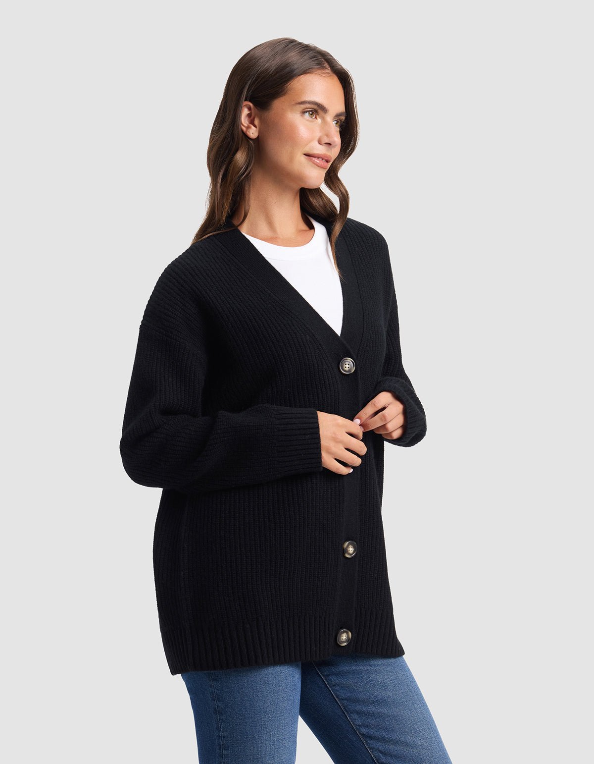 A woman stands against a plain gray background, looking to the side and wearing Cozy Earth's Oversized Sunday Cashmere Cardigan in black over a white top and blue jeans.