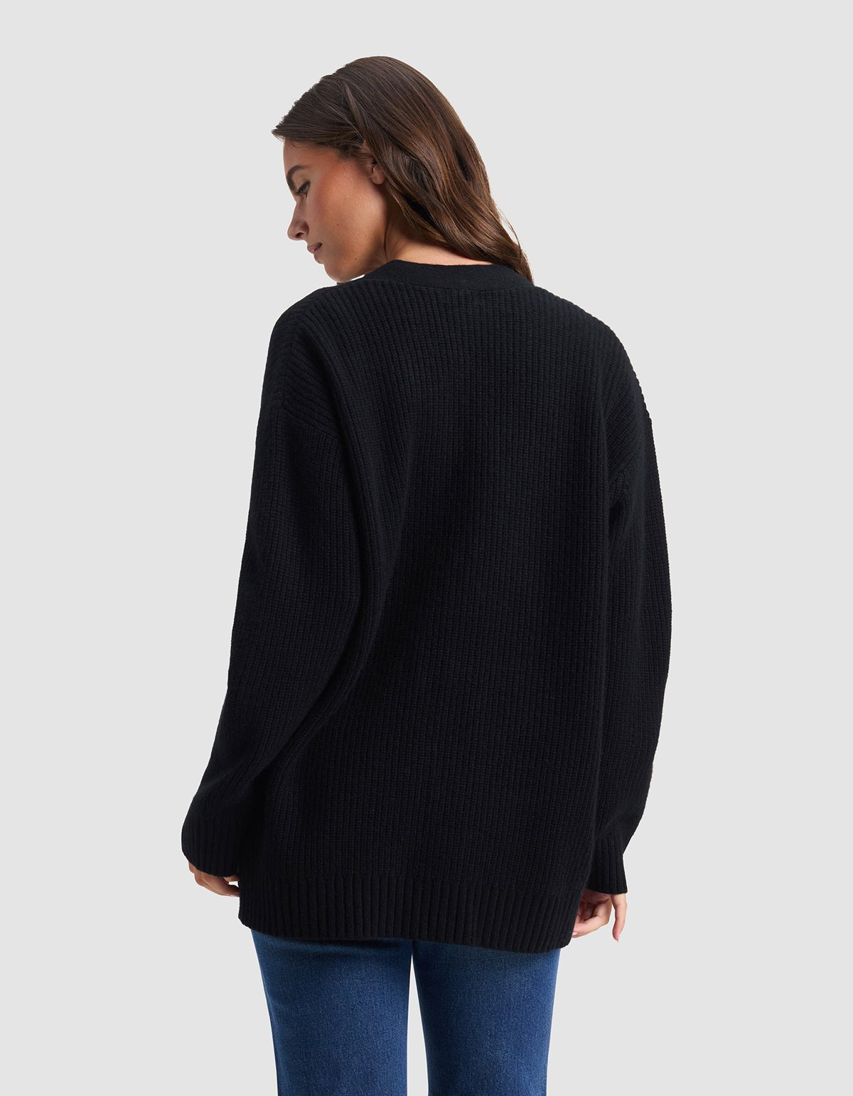 A woman with long brown hair models the Cozy Earth Oversized Sunday Cashmere Cardigan, standing in blue jeans and looking over her shoulder against a plain light gray background.