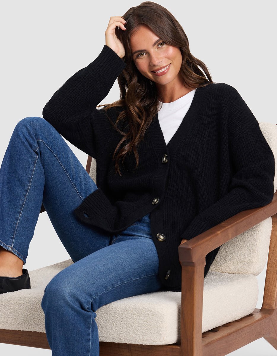 A woman with long brown hair smiles as she sits comfortably on a cushioned wooden chair, wearing the Cozy Earth Oversized Sunday Cashmere Cardigan over a white shirt and blue jeans, her hand resting on her head.