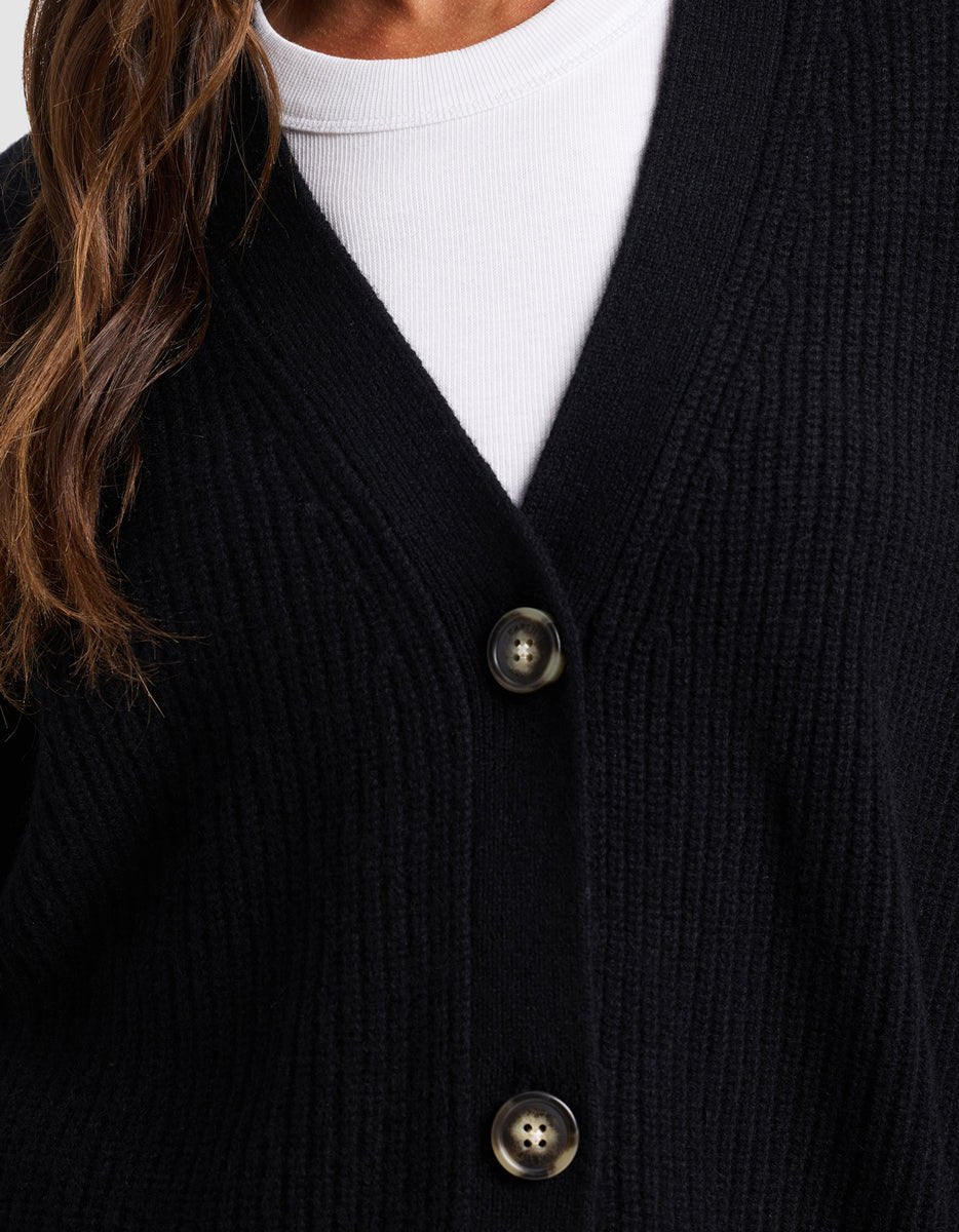 Close-up of a person wearing Cozy Earth's black Oversized Sunday Cashmere Cardigan, featuring large buttons and a ribbed knit, layered over a white crew-neck shirt. Only part of the person's face and hair appears on the left side.