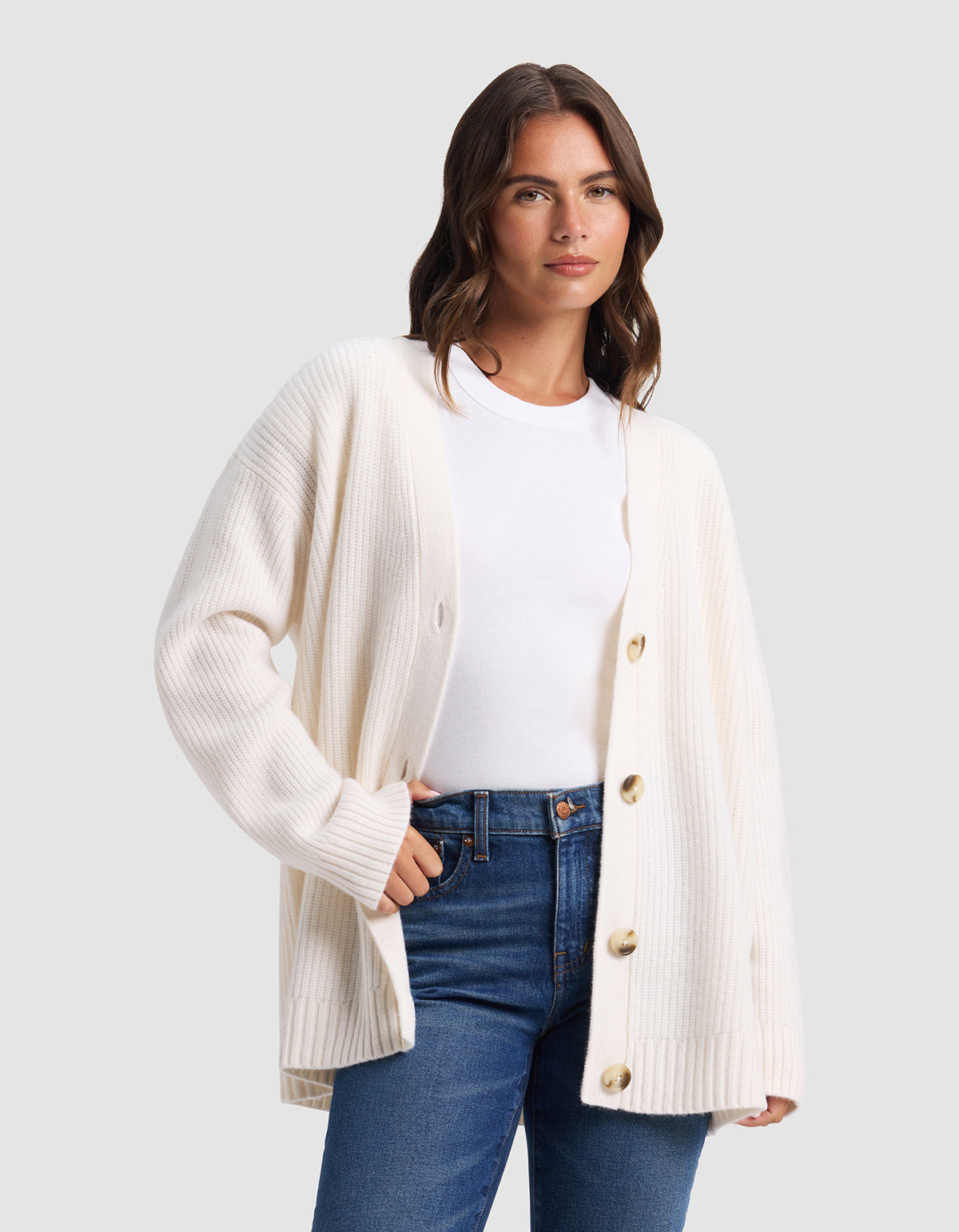 A woman stands against a plain light grey background wearing the Cozy Earth Oversized Sunday Cashmere Cardigan in white, styled over a white t-shirt and blue jeans, with one hand casually in her pocket.|Color:Bone