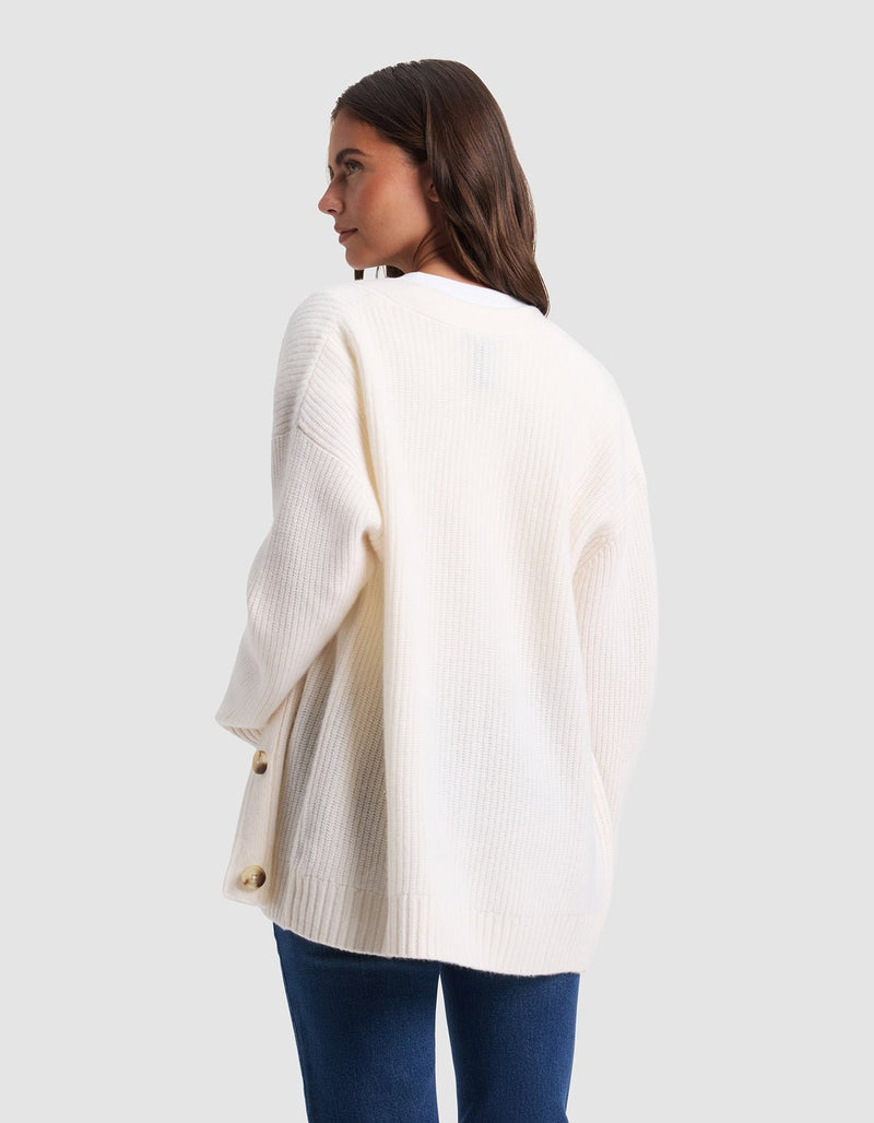 A woman with long brown hair, dressed in blue jeans, stands facing away against a plain light gray background wearing Cozy Earth's Oversized Sunday Cashmere Cardigan—a loose, white, ribbed design with large side buttons.