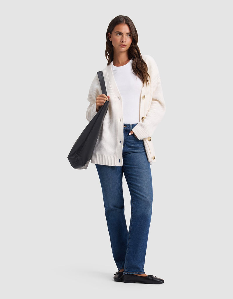 A woman stands against a light gray background, wearing the Cozy Earth Oversized Sunday Cashmere Cardigan in white over a white shirt, blue jeans, and black shoes. She carries a black tote bag on her shoulder and looks ahead with a neutral expression.