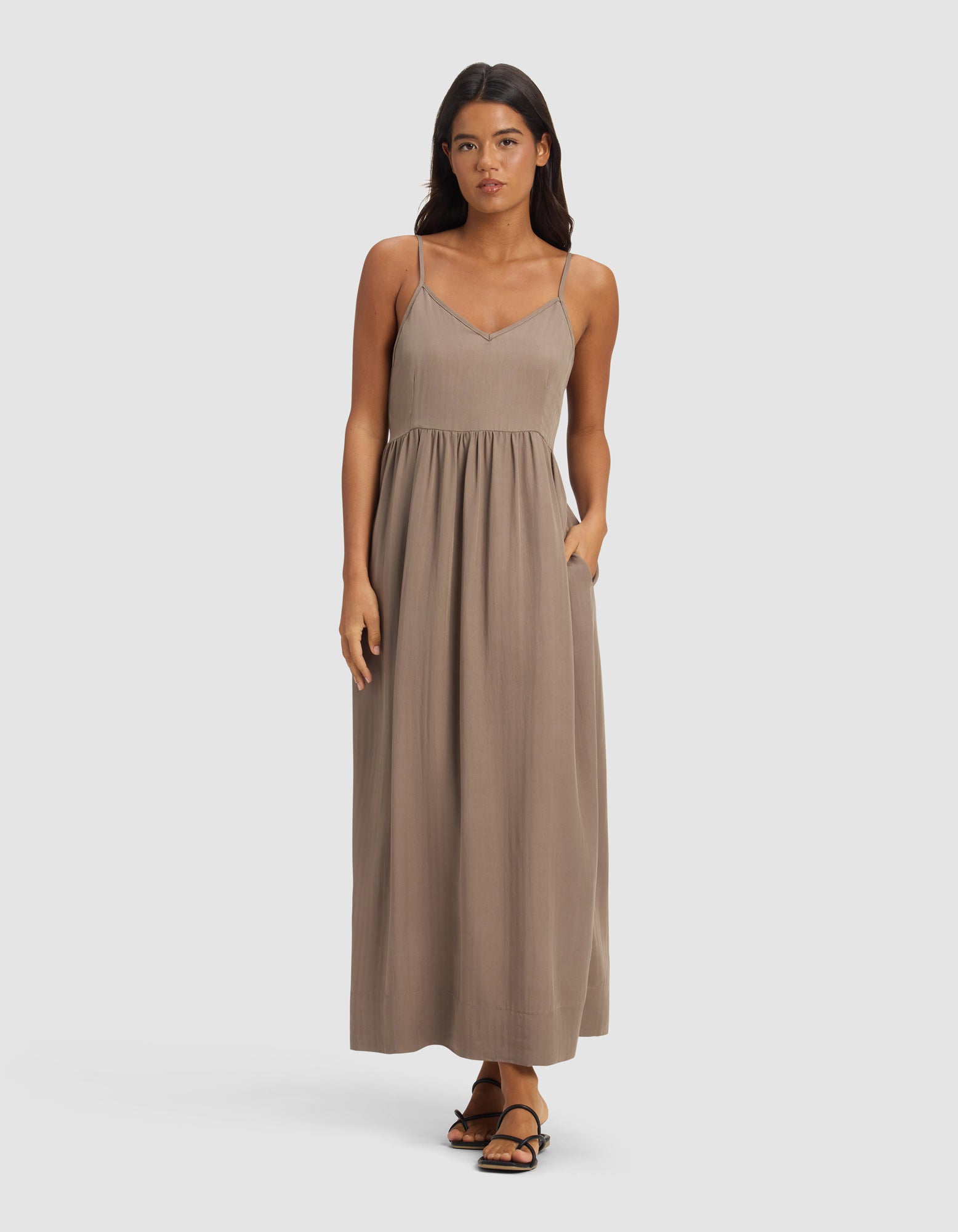 A woman models the Cozy Earth Women's Sunset Gathered Dress, an ankle-length, sleeveless taupe dress with thin straps and a gathered waist. She wears black sandals and poses with one hand in her pocket against a light background.|Color:Ebony