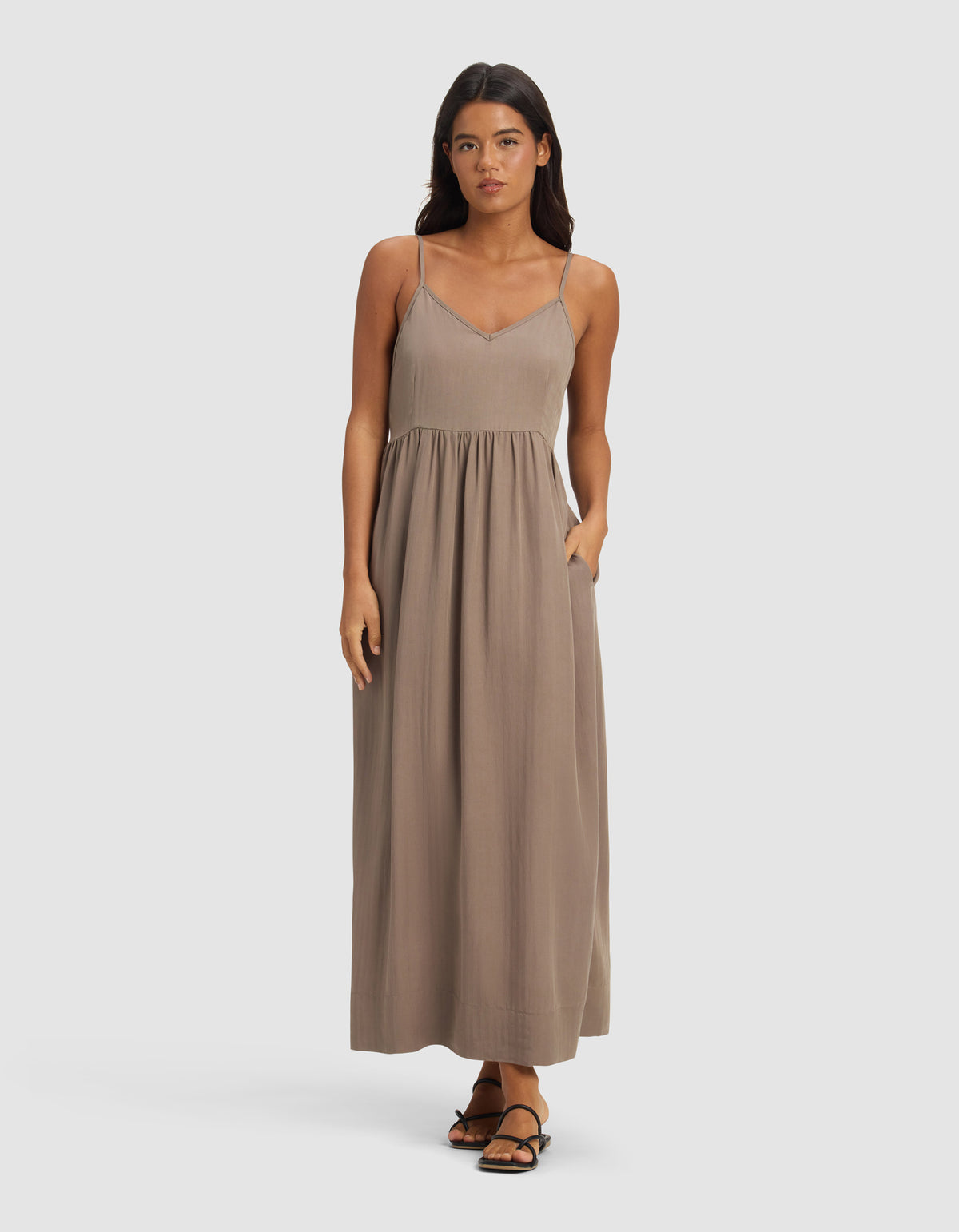 A woman faces forward wearing Cozy Earth's Women's Sunset Gathered Dress - Last Chance, a sleeveless, loose taupe maxi dress with thin straps. She has long dark hair, her hands by her sides, and stands against a plain light background.