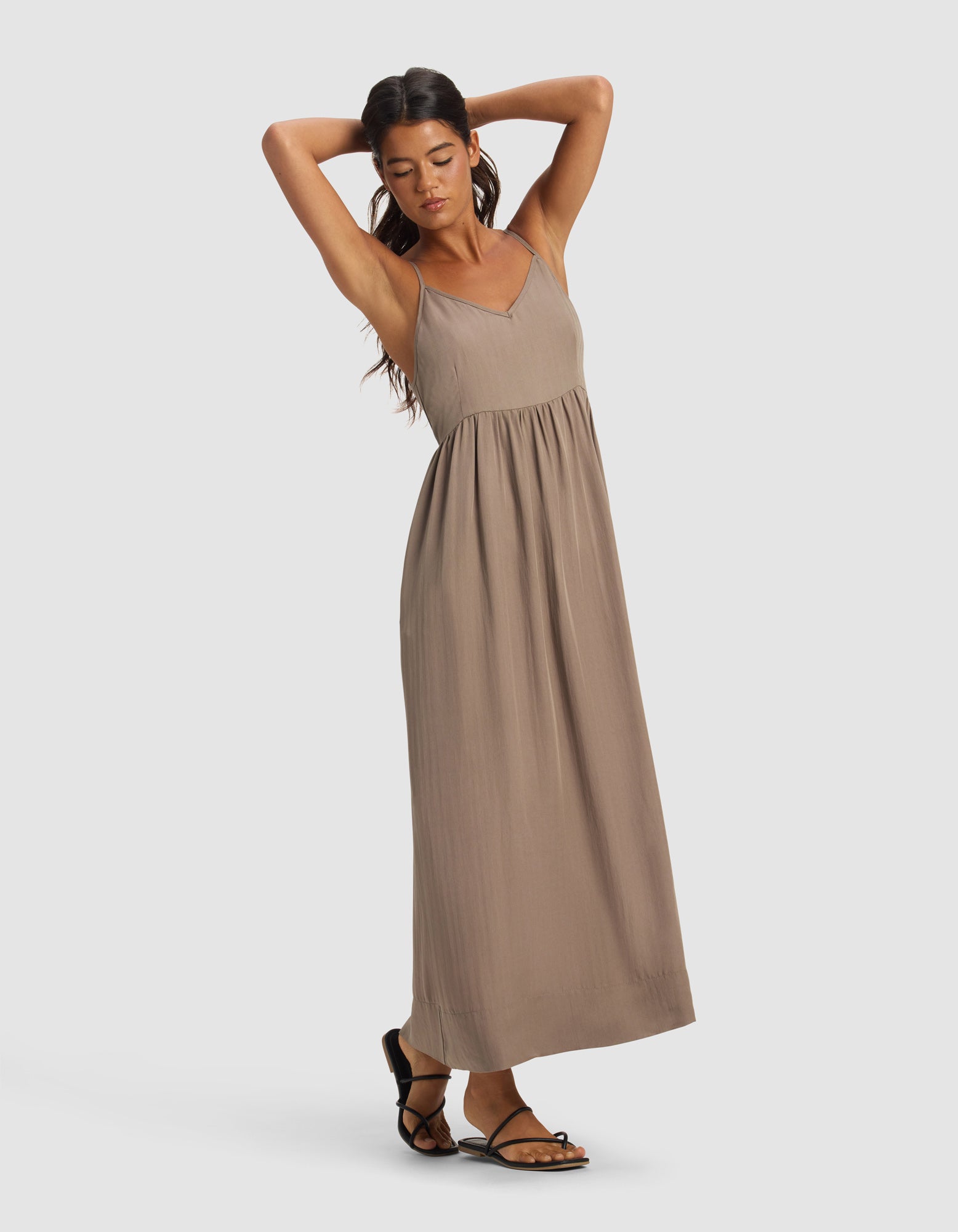 A woman models the Cozy Earth Women's Sunset Gathered Dress - Last Chance in taupe, a sleeveless maxi with thin straps. She stands barefoot against a plain background, arms raised and one leg slightly bent.