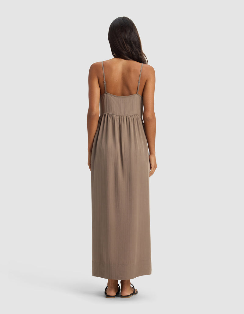 A woman with long dark hair, seen from behind, wears the Cozy Earth Women's Sunset Gathered Dress—sleeveless, ankle-length with thin straps—paired with flat sandals, standing against a plain light gray background.