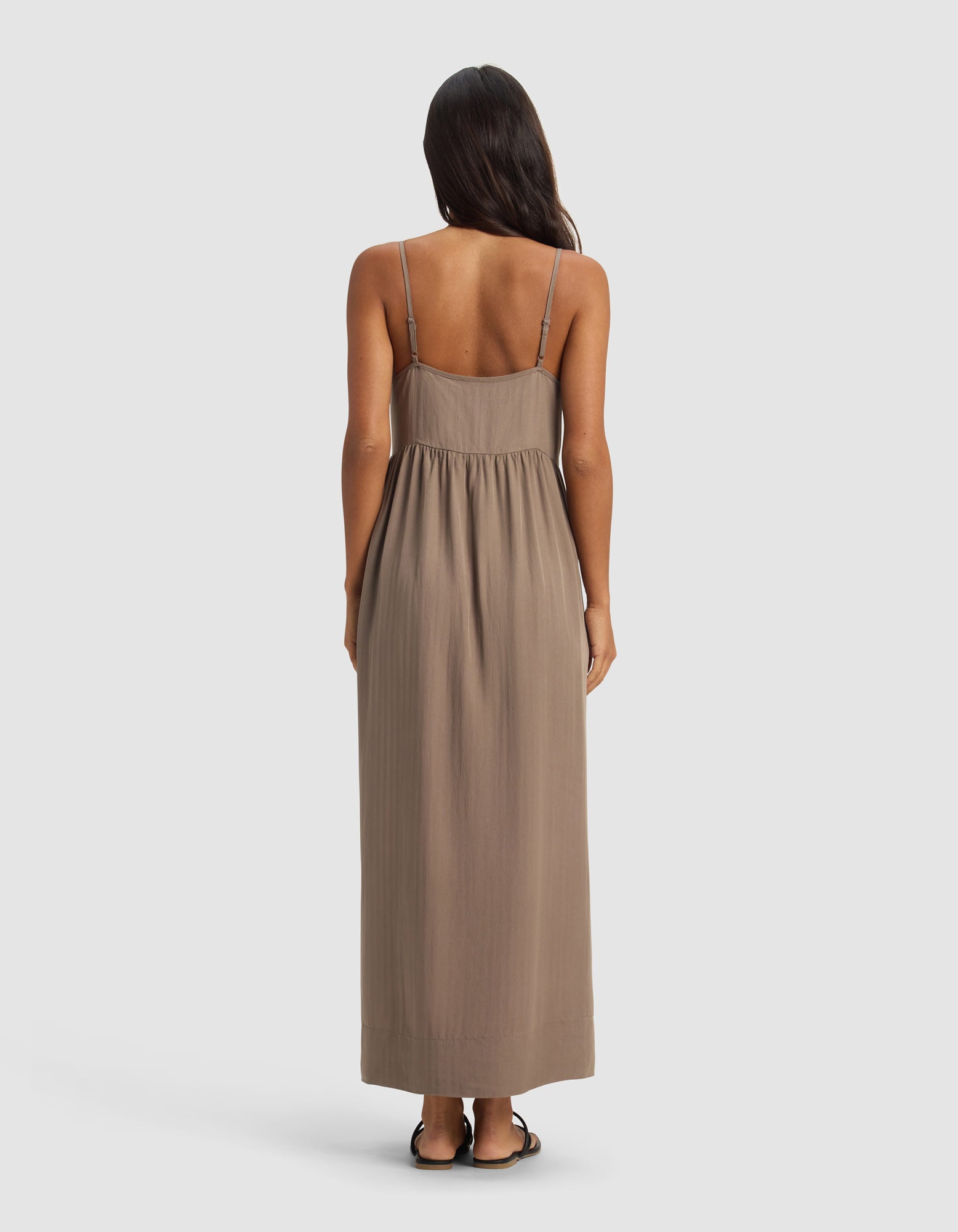 A woman with long dark hair faces away, wearing Cozy Earth's Women's Sunset Gathered Dress – Last Chance, a sleeveless, ankle-length taupe style with thin straps and flat sandals, set against a plain light background.