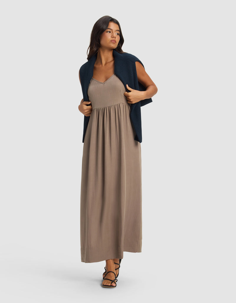 A woman models the Cozy Earth Women's Sunset Gathered Dress in taupe, styled with black sandals and a navy sweater over her shoulders, standing against a plain light gray background and looking slightly to the side.