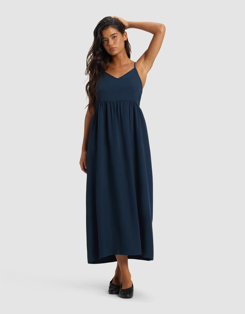 A woman poses on a plain background wearing Cozy Earth's Women's Sunset Gathered Dress - Last Chance, a sleeveless navy maxi with a V-neck and empire waist. She has long, dark hair and black shoes, touching her head with one hand.