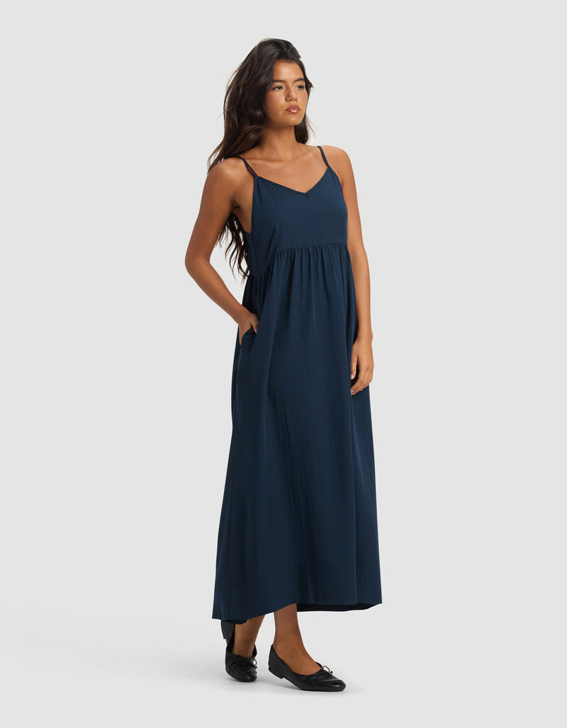 A woman with long dark hair stands against a plain background wearing Cozy Earth's Women's Sunset Gathered Dress - Last Chance, a sleeveless navy blue, ankle-length dress with thin straps and black flats, one hand in her pocket.