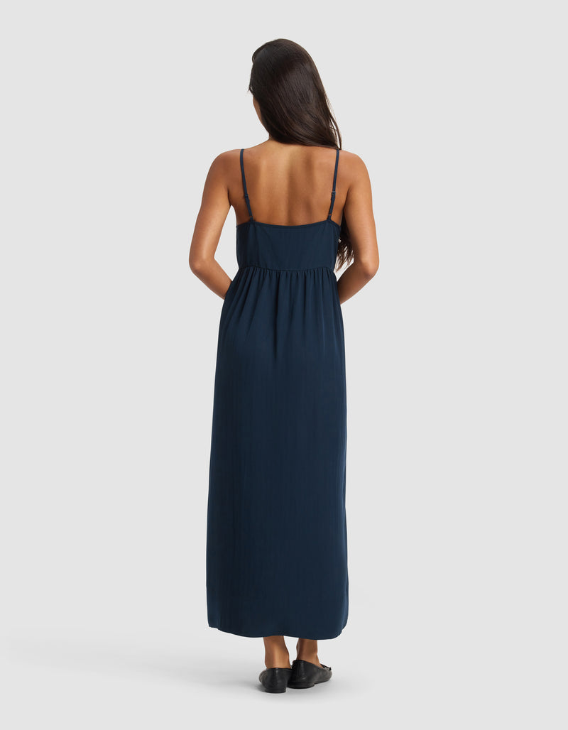 A woman with long dark hair stands facing away, wearing the Cozy Earth Women's Sunset Gathered Dress in navy blue with thin straps and black shoes, set against a plain white background.