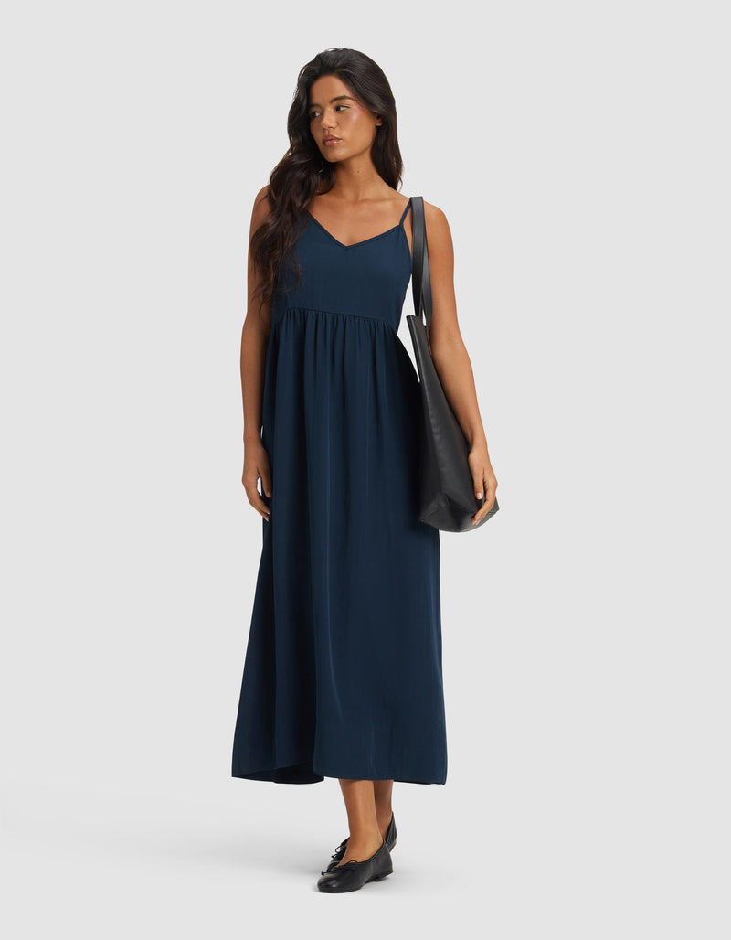 A woman in the Cozy Earth Women's Sunset Gathered Dress - Last Chance, a sleeveless navy maxi with a V-neckline, styled with black flats and a large black shoulder bag, stands against a plain light gray background.