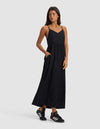 A woman wearing the Cozy Earth Women's Sunset Gathered Dress - Last Chance stands against a plain light gray background, looking to her left with one hand in her pocket and pairing the dress with black Adidas sneakers.|Color:Jet Black
