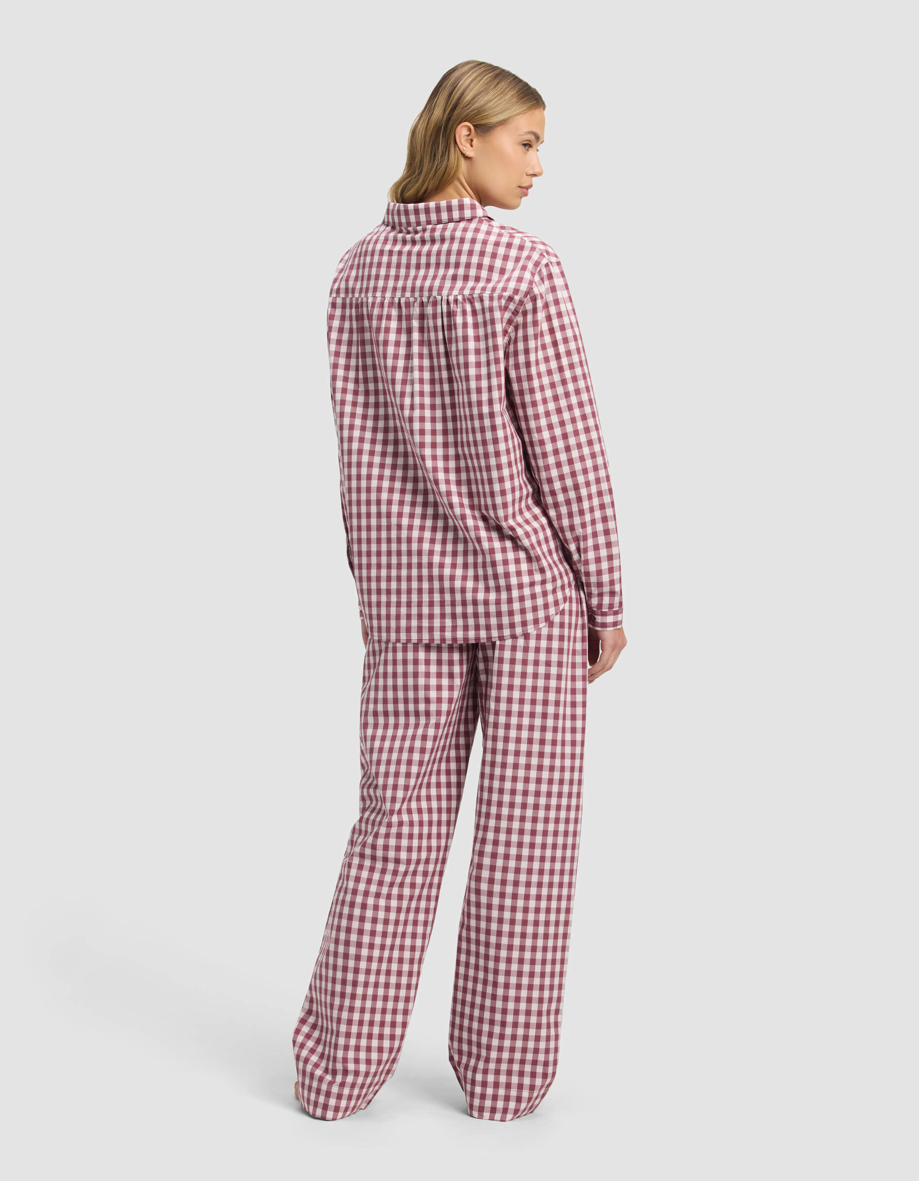 A person stands facing away, wearing the Cozy Earth Women's Sutton Long Sleeve Pajama Set in a red and white checkered pattern. The background is plain light gray.|Color:Mulberry Gingham