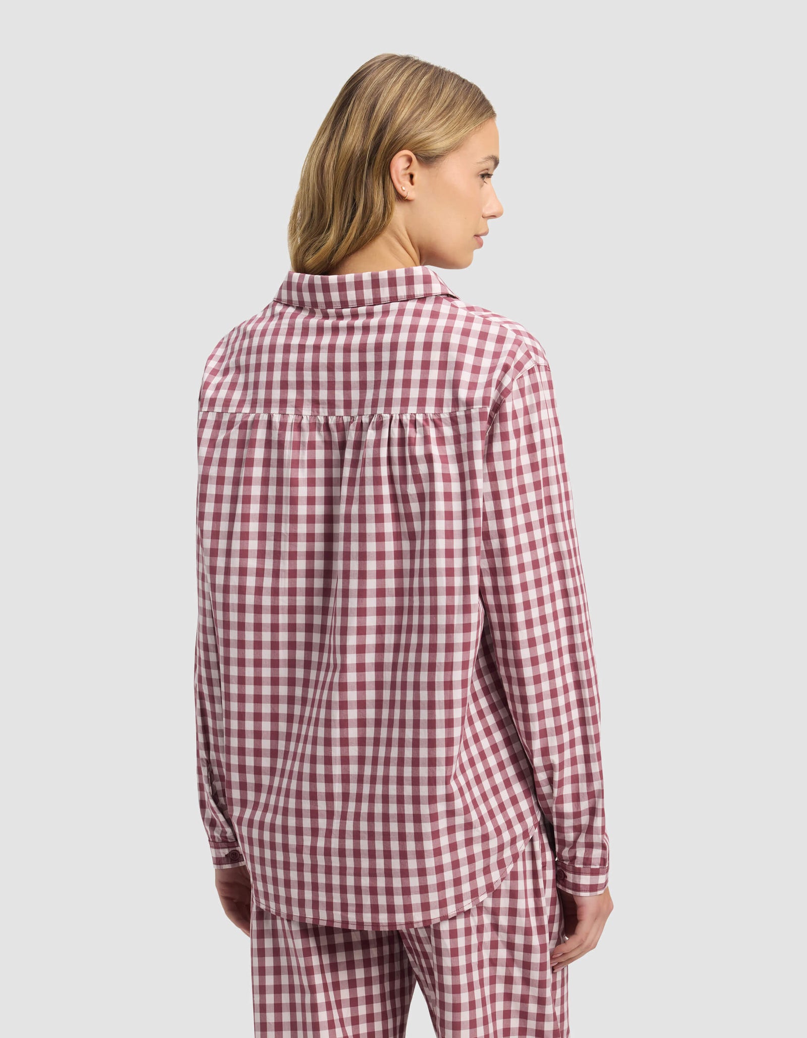 A woman with blonde hair wears the Cozy Earth Women's Sutton Long Sleeve Pajama Top in a red and white check, with matching pants, standing facing away against a plain light gray background.