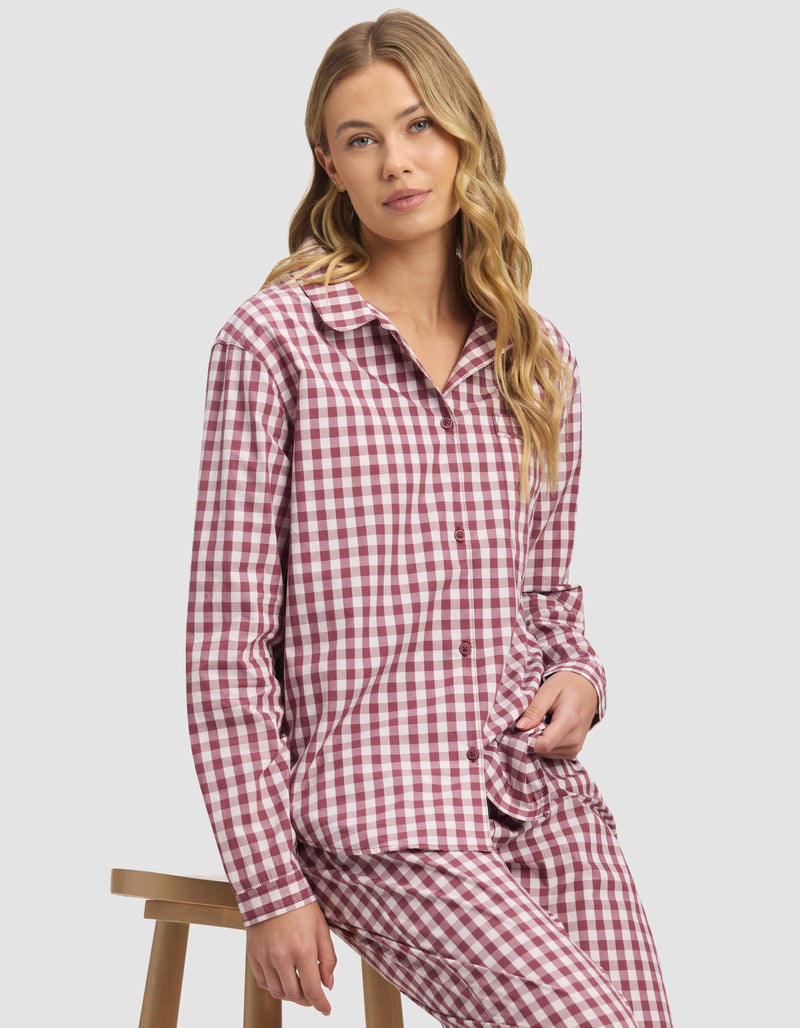 A woman with long blonde hair sits on a wooden stool, wearing the Cozy Earth Women's Sutton Long Sleeve Pajama Top in red and white checks, paired with matching pants, and looks to the side with a neutral expression.