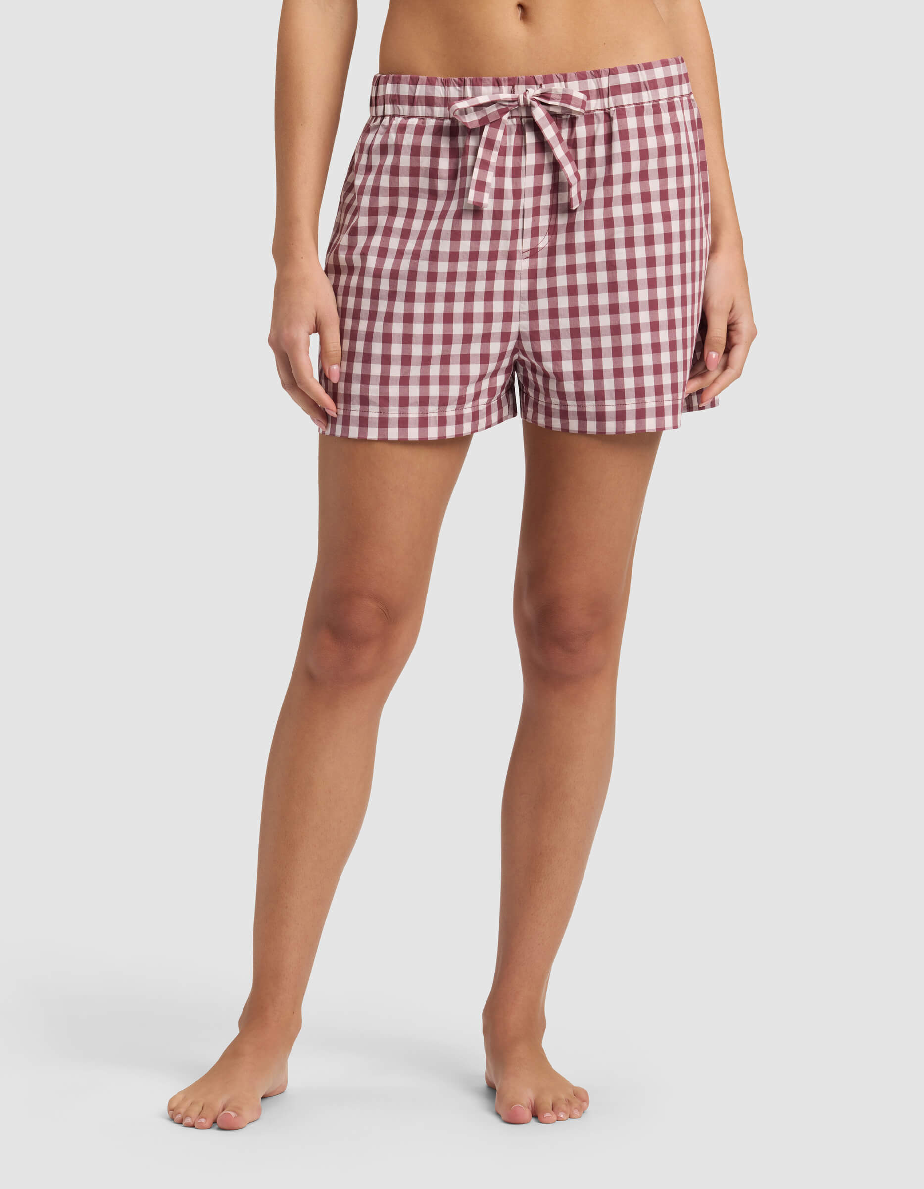 A model wears Cozy Earth's Women's Sutton Pajama Short—red and white gingham with a drawstring waist, barefoot and shown from waist to feet against a plain white background.