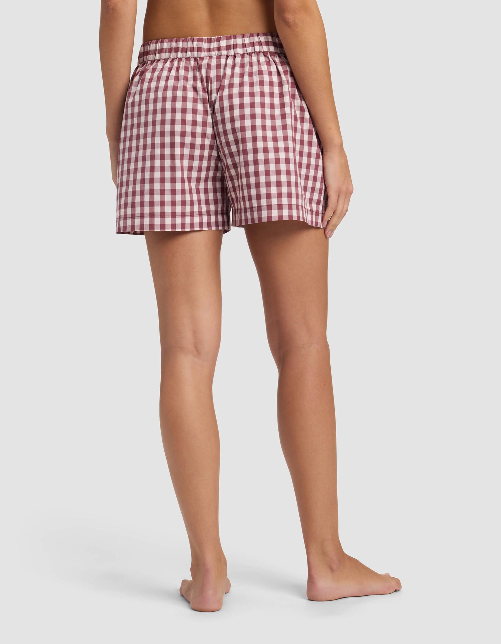 A person stands barefoot, facing away, wearing Cozy Earth's Women's Sutton Pajama Short in a red and white checkered pattern with an elastic waistband, against a plain white background.