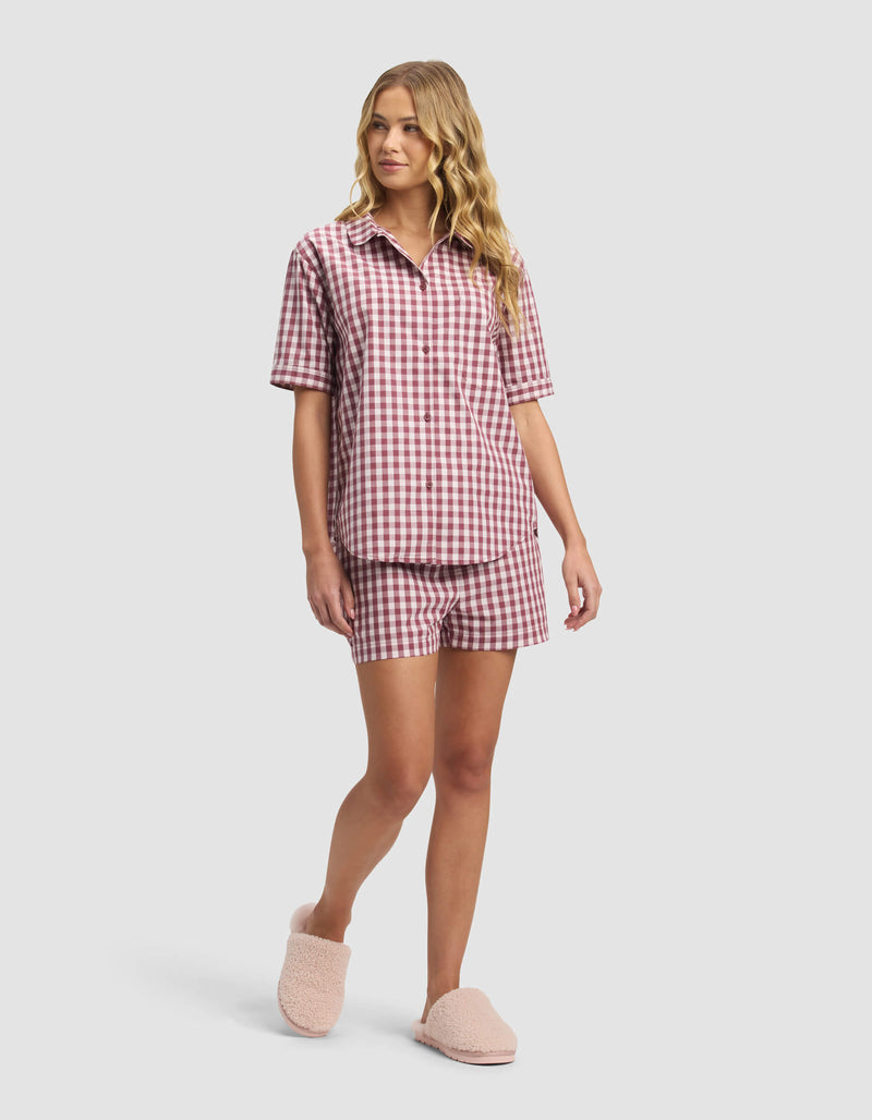 A woman with long blonde hair models the Cozy Earth Women's Sutton Pajama Short set—matching red and white gingham short-sleeve button-up shirt and shorts—paired with light pink fuzzy slippers, standing before a plain light gray background.