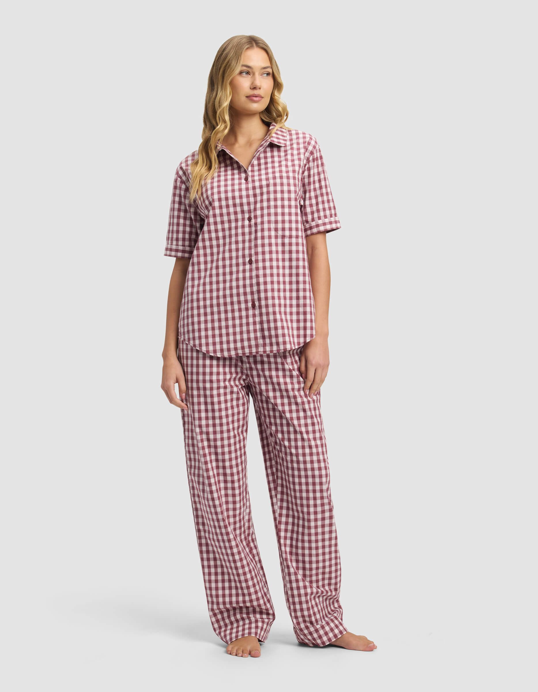 A woman with long, wavy blond hair wears the Cozy Earth Women's Sutton Short Sleeve & Pant Pajama Set featuring a red and white checkered pattern, including a button-up shirt and loose-fitting pants. She stands barefoot with a neutral expression.|Color:Mulberry Gingham