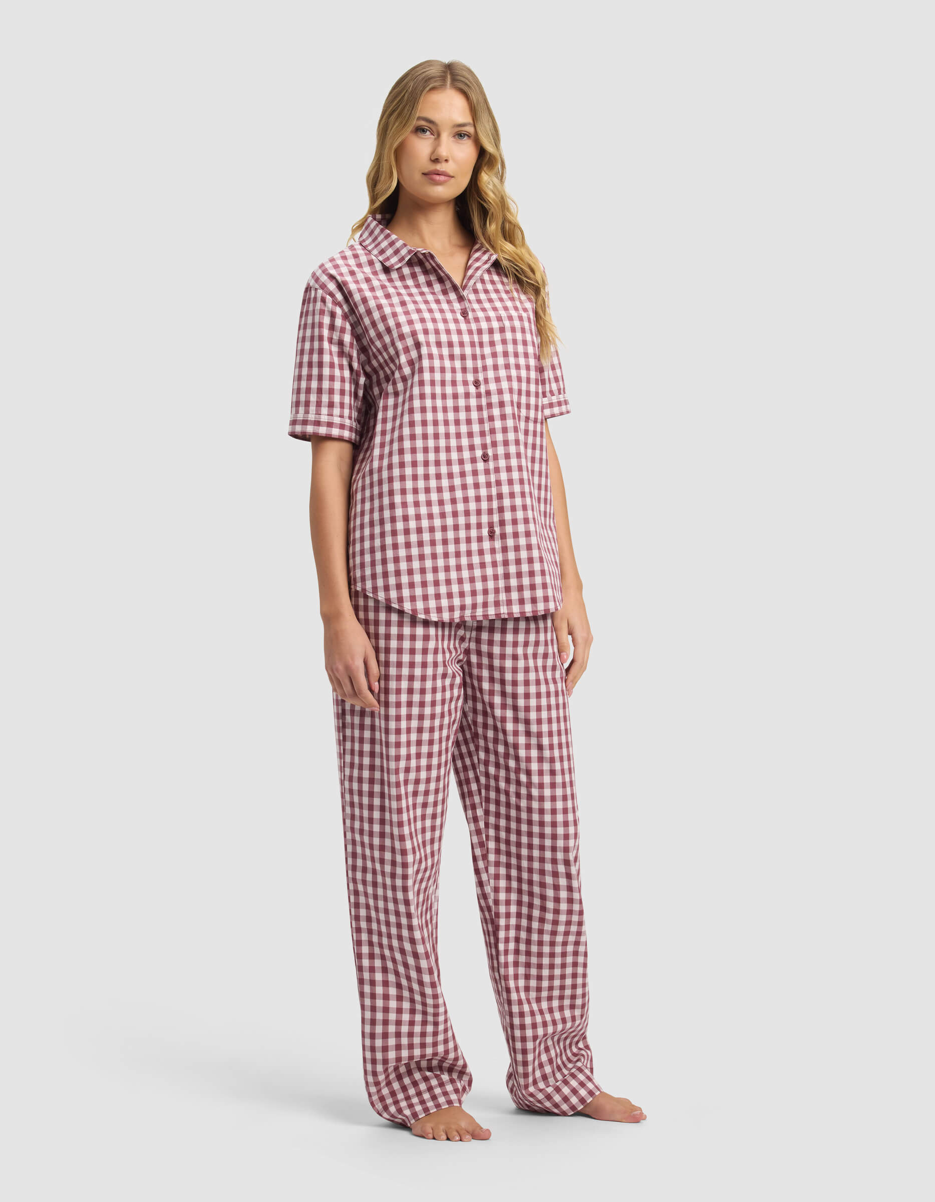 A woman stands barefoot in the Cozy Earth Women's Sutton Short Sleeve & Pant Pajama Set, featuring a red and white checkered pattern. The matching set has a button-up shirt and loose pants, shown against a plain light background.|Color:Mulberry Gingham
