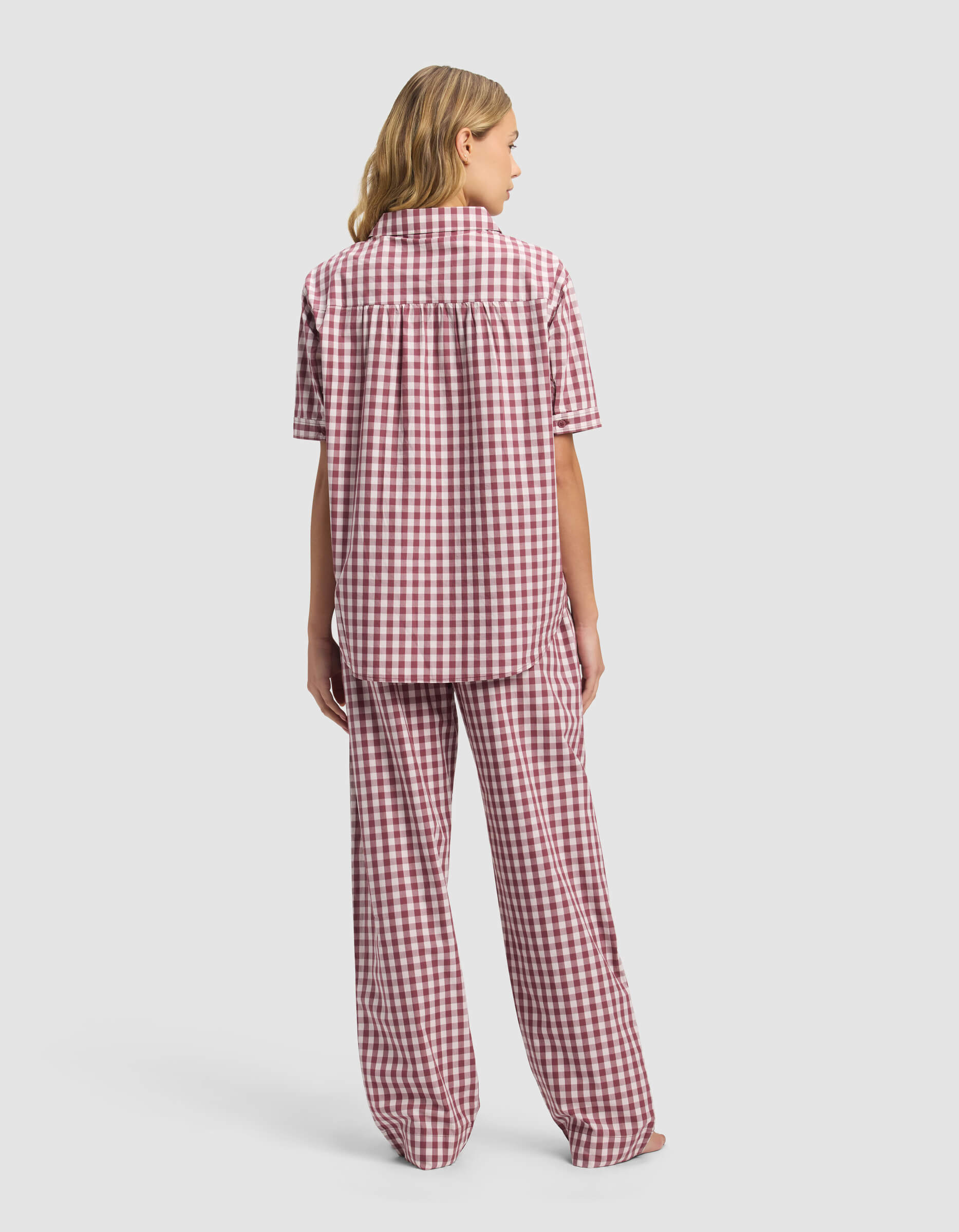 A person with blonde hair stands facing away, wearing the Cozy Earth Women's Sutton Short Sleeve & Pant Pajama Set in a red and white checkered pattern, set against a plain light gray background.|Color:Mulberry Gingham