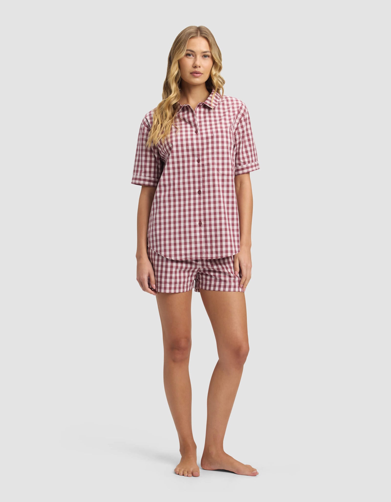 A woman stands barefoot against a plain light background wearing the Cozy Earth Women's Sutton Short Sleeve Pajama Set, featuring a red and white checkered pattern with a button-up shirt and matching shorts.