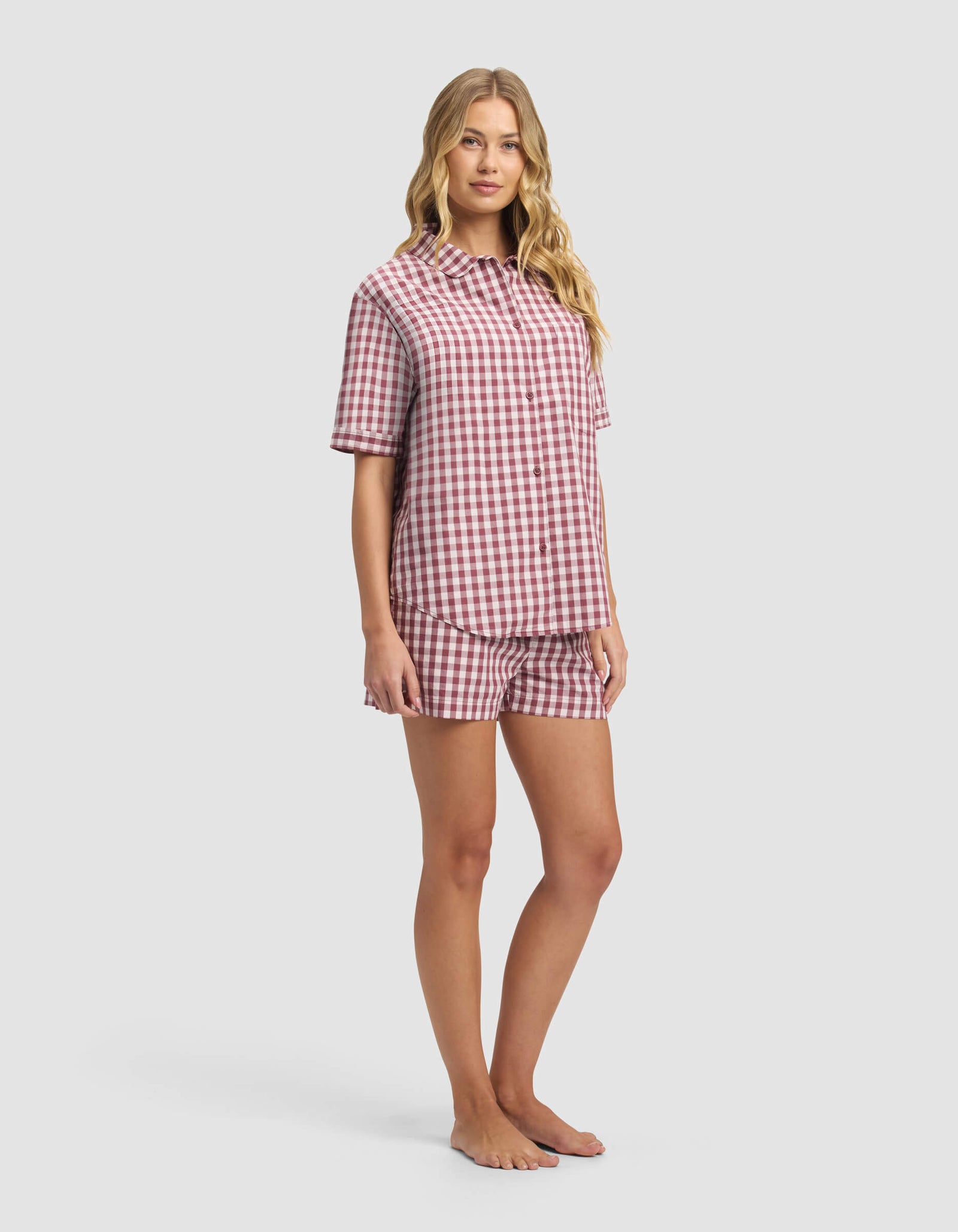 A woman stands barefoot against a light gray background, wearing Cozy Earth's Women's Sutton Short Sleeve Pajama Set in a red and white gingham pattern.