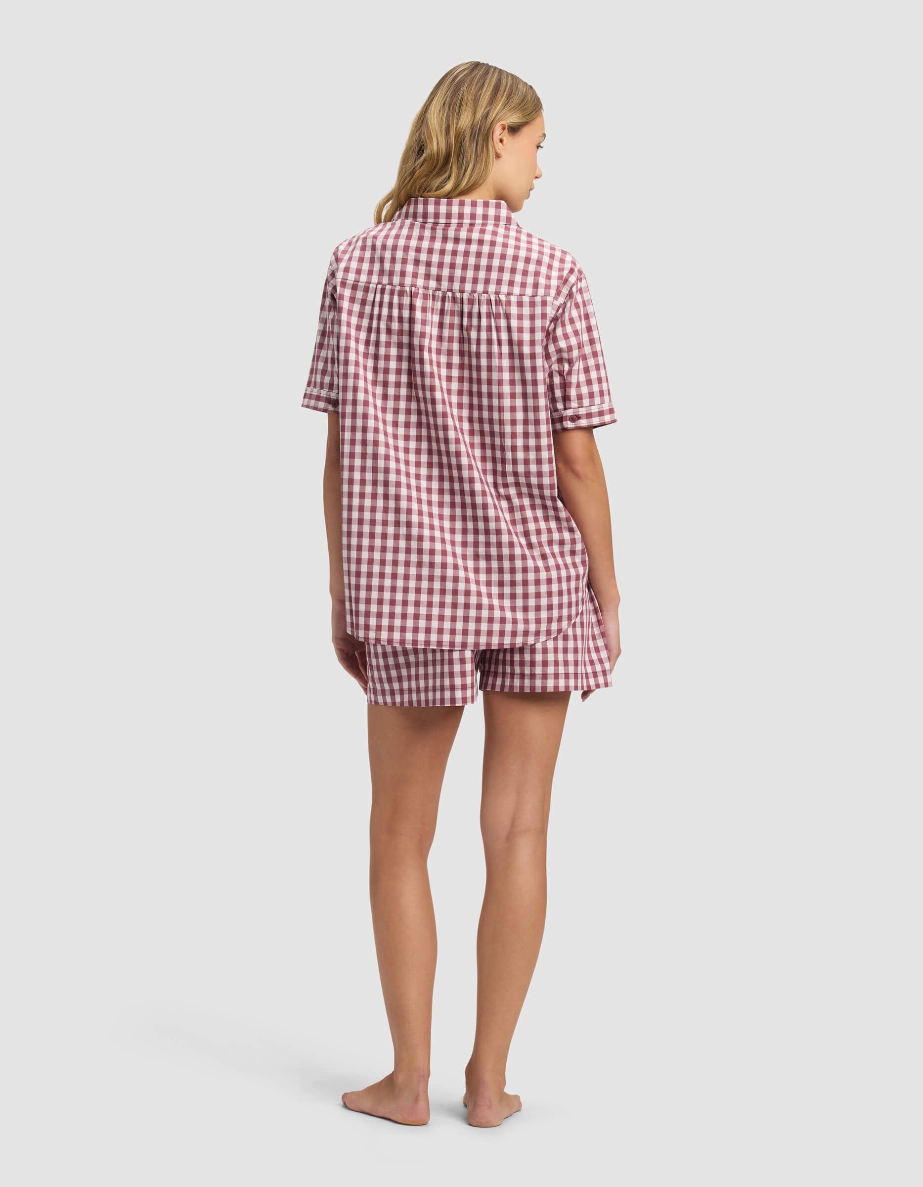 A barefoot blonde woman faces away, wearing Cozy Earth's Women's Sutton Short Sleeve Pajama Set— a loose red and white checkered shirt with matching shorts—against a plain white background.|Color:Mulberry Gingham