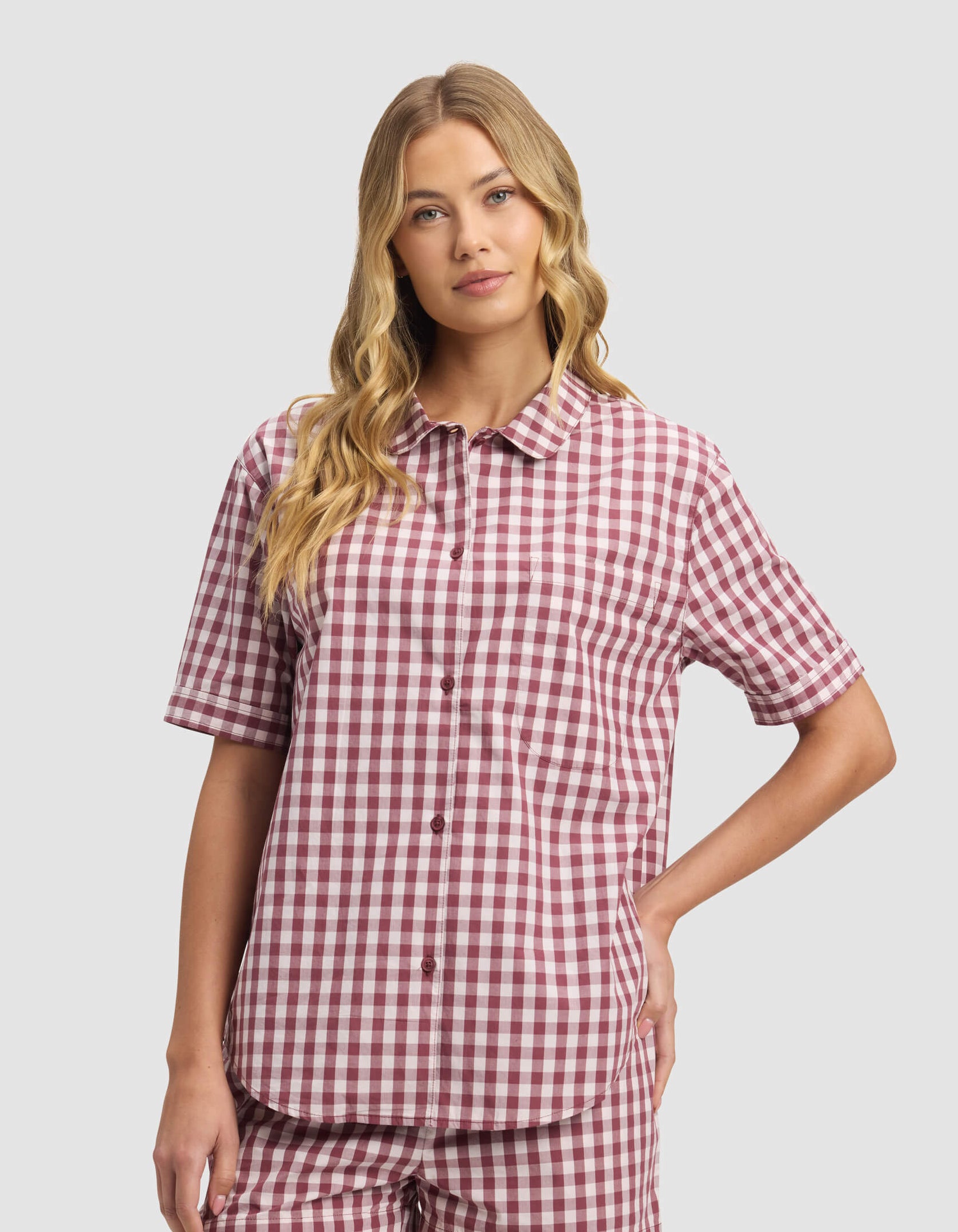 A woman with long blonde hair wears the Cozy Earth Women's Sutton Short Sleeve Pajama Top in a red and white gingham pattern, paired with matching shorts, standing against a plain light gray background.