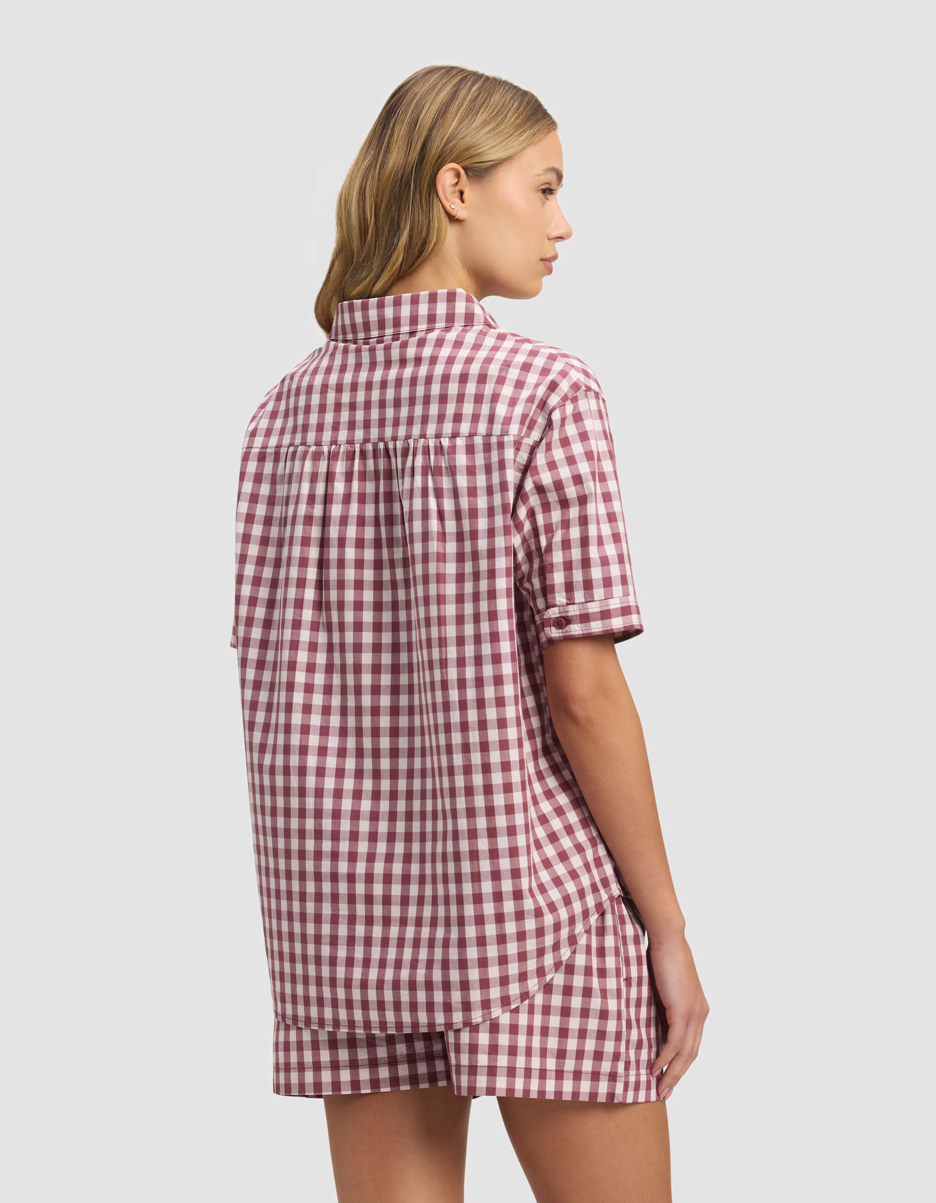 A woman with light brown hair, seen from behind against a plain light background, wears the Cozy Earth Women's Sutton Short Sleeve Pajama Top in an oversized red and white gingham pattern with matching shorts.|Color:Mulberry Gingham