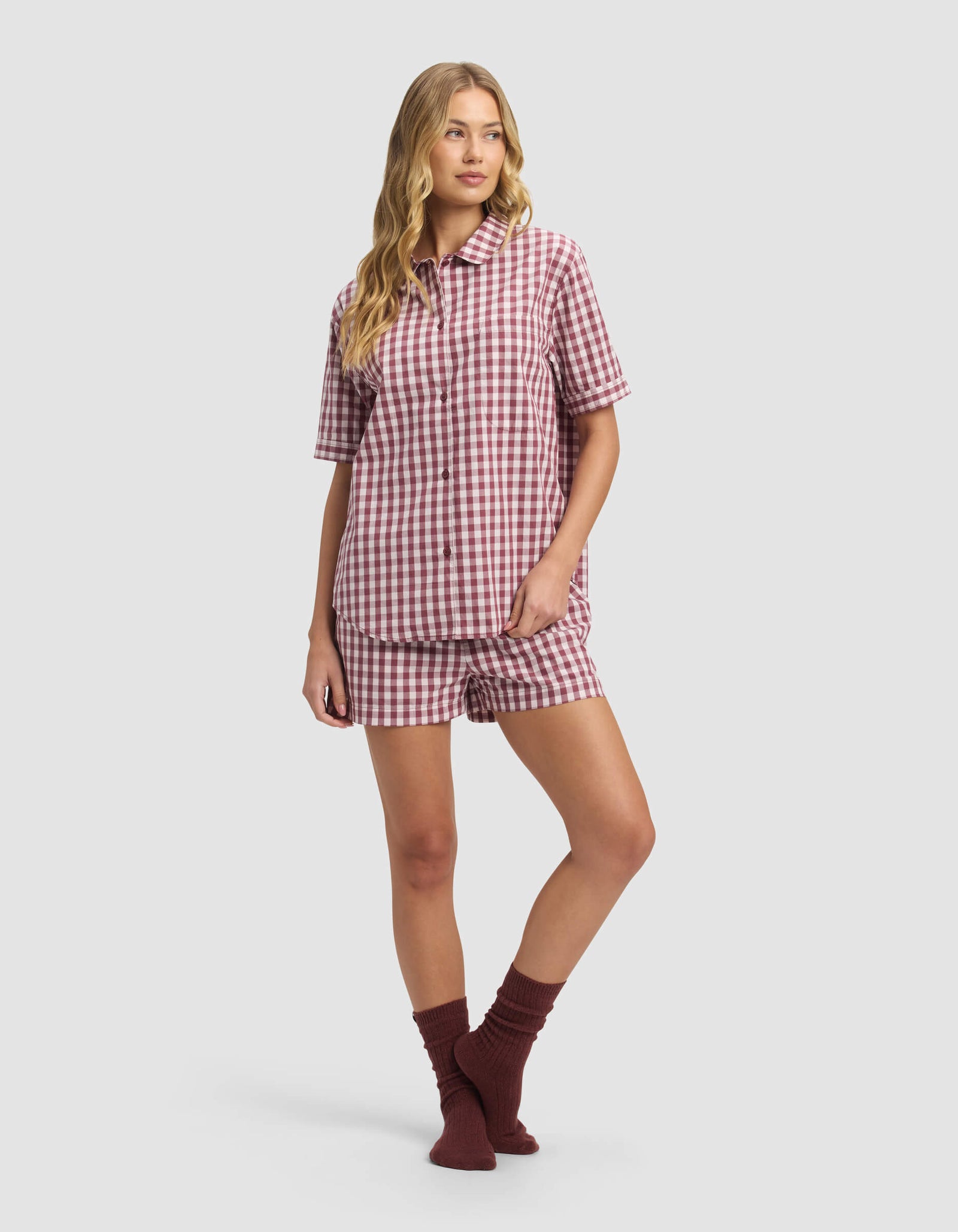 A person wears the Cozy Earth Women's Sutton Short Sleeve Pajama Top in red and white gingham with matching shorts and dark red socks, standing against a plain light gray background.
