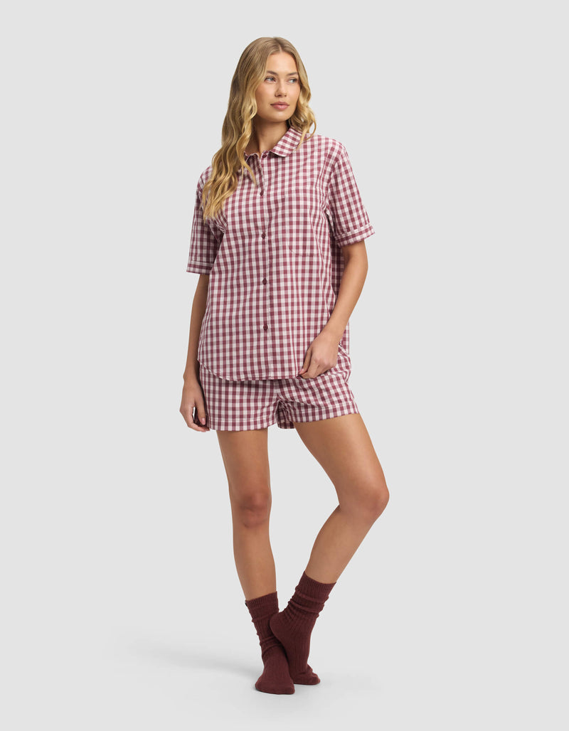 A person wears the Cozy Earth Women's Sutton Short Sleeve Pajama Top in red and white gingham with matching shorts and dark red socks, standing against a plain light gray background.