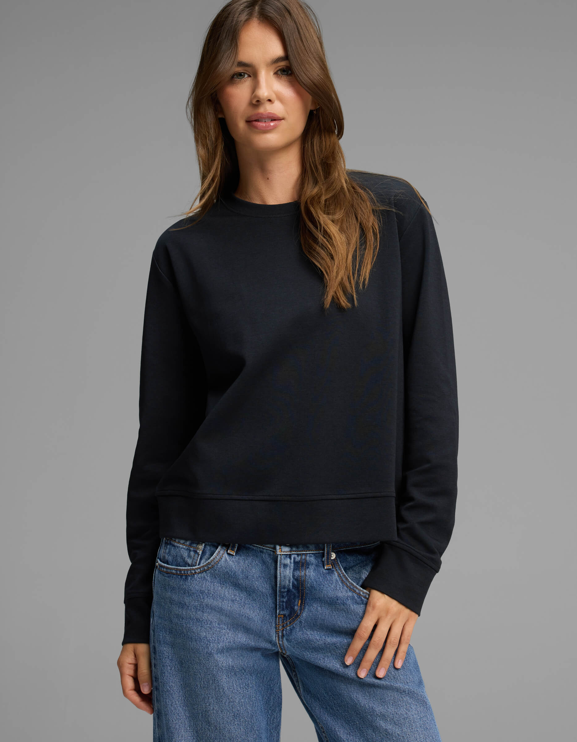 A woman with long brown hair stands against a plain gray background, looking at the camera with a slight smile while wearing Cozy Earth's Women's Versa Crewneck in black and blue jeans.
