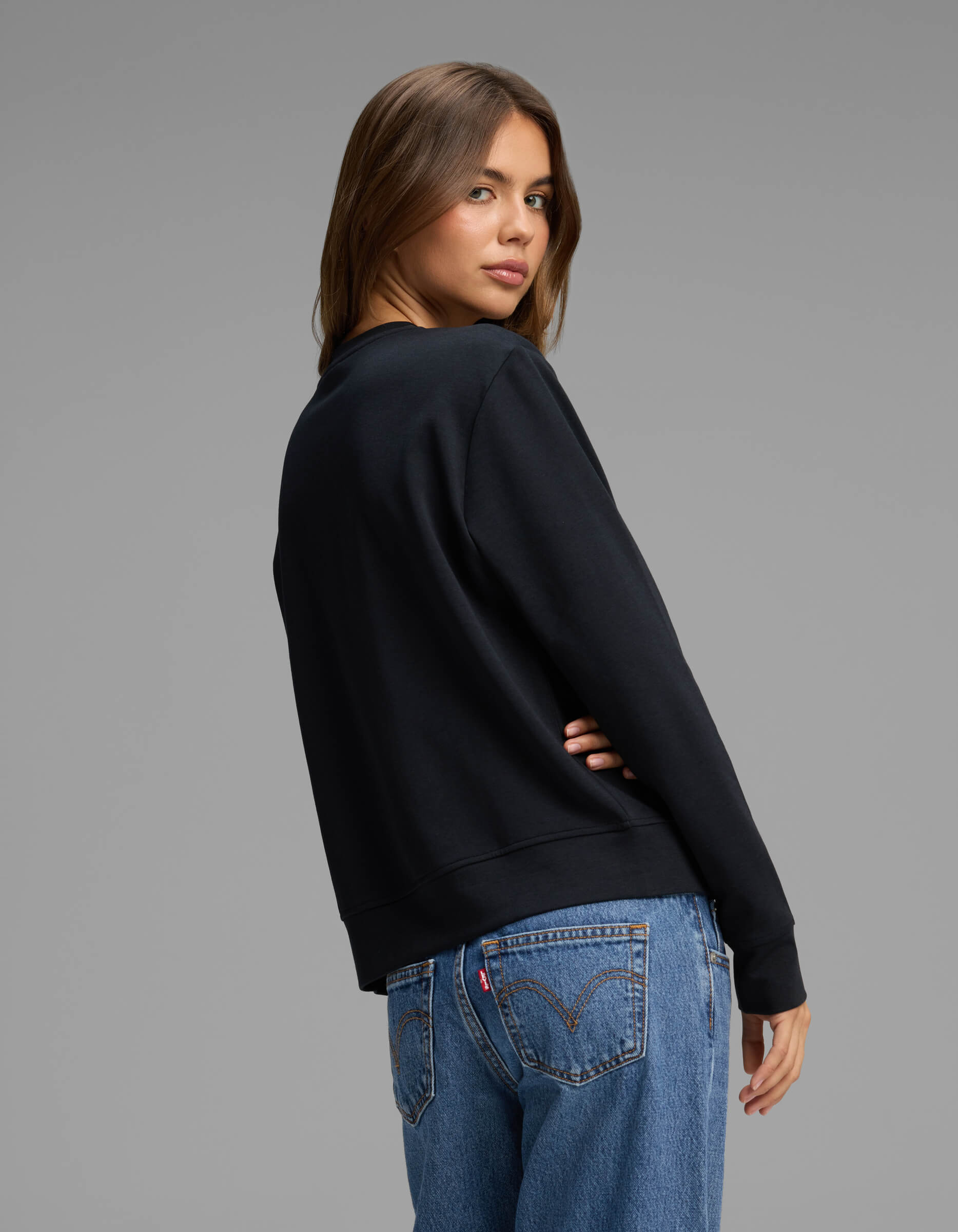 A woman with long brown hair wears the Cozy Earth Women's Versa Crewneck in black and blue jeans, standing with her back turned and head looking over her shoulder against a plain gray background.