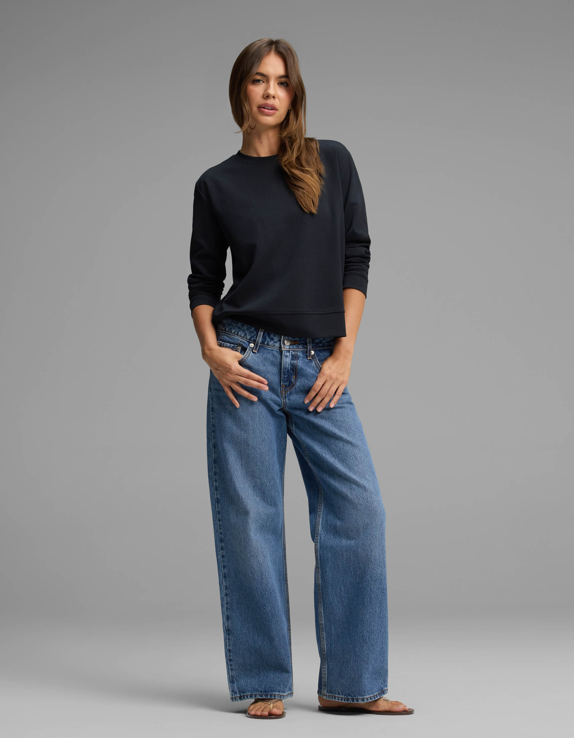 A woman with long brown hair stands against a gray background wearing the Cozy Earth Women's Versa Crewneck, paired with loose blue jeans and sandals. She poses with one hand in her pocket and the other resting by her side.