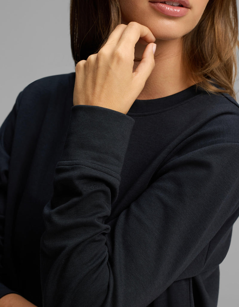 A woman wearing the Cozy Earth Women's Versa Crewneck in black rests her chin on her hand, with only the lower half of her face and upper torso visible.