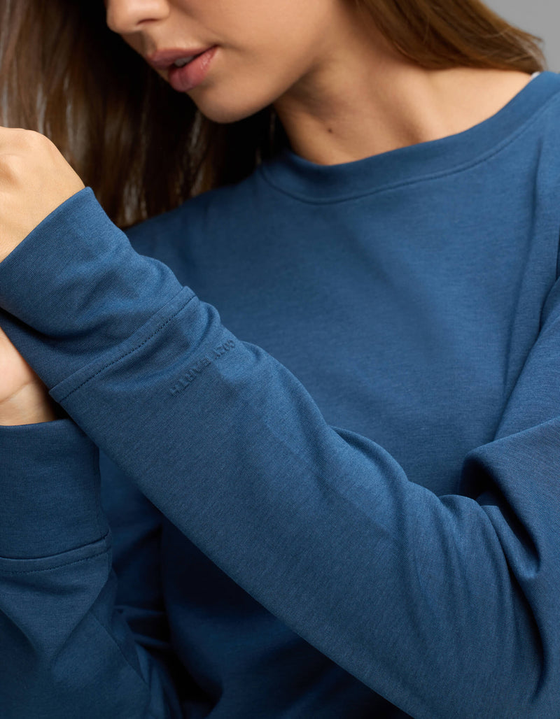 A woman wearing the Cozy Earth Women's Versa Crewneck in blue, featuring subtle embroidered text on the sleeve, holds her hands together near her face. Her head is turned slightly to the side, and only part of her face is visible.