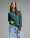 A woman with long brown hair wears the Cozy Earth Women's Versa Crewneck in green and light blue jeans, posing with one hand in her pocket against a plain gray background.|Color:Uniform