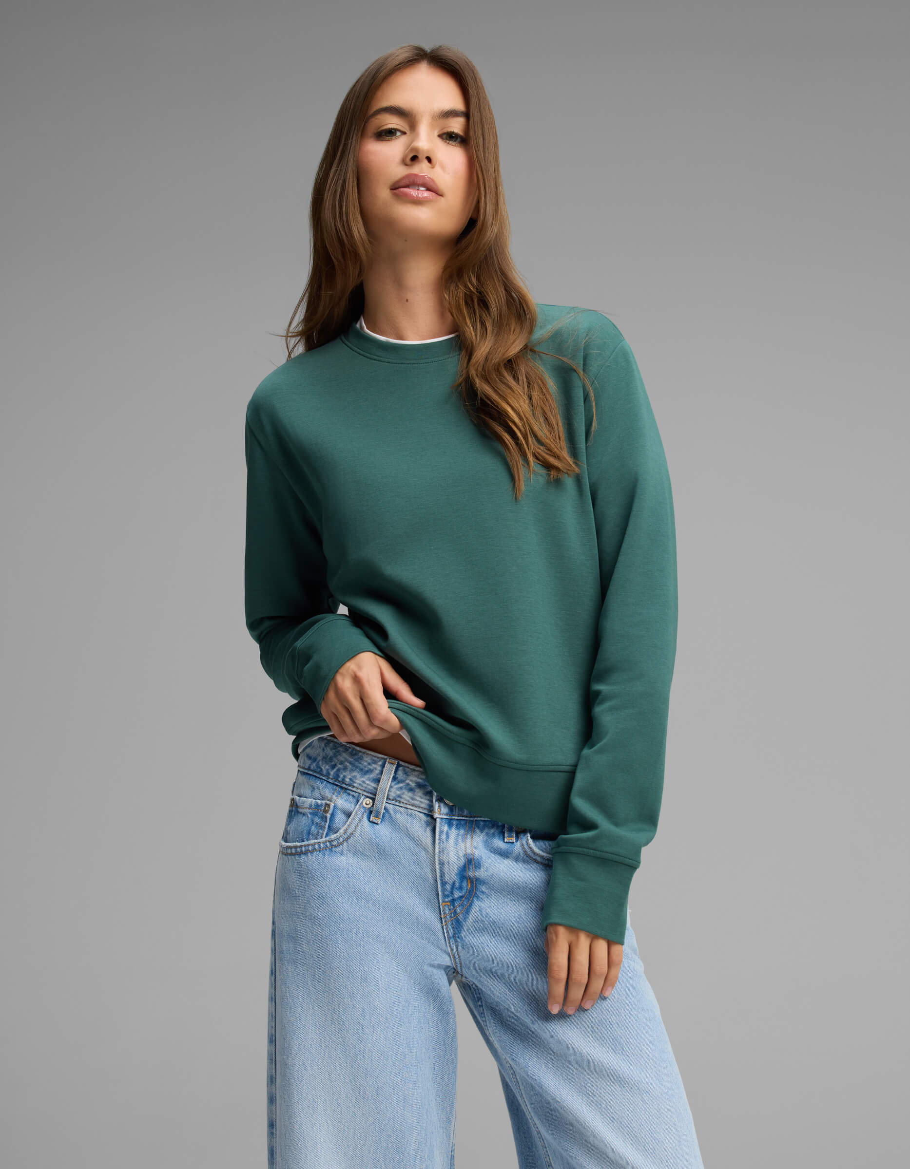 A woman with long brown hair wears the Cozy Earth Women's Versa Crewneck in green and light blue jeans, posing with one hand in her pocket against a plain gray background.|Color:Uniform