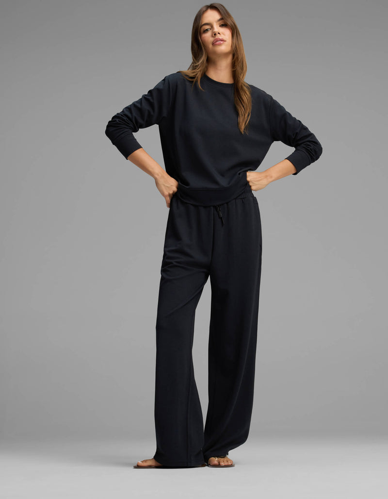 A woman stands against a gray background wearing the Cozy Earth Women's Versa Crewneck & Straight Leg Pant Set in black, paired with sandals. She has long brown hair and poses confidently with her hands on her hips, looking at the camera.
