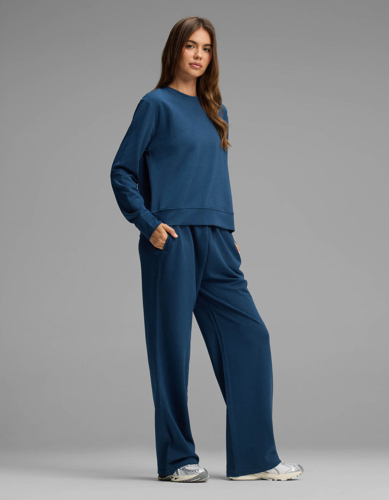 A person stands against a plain gray background, wearing the Cozy Earth Women's Versa Crewneck & Straight Leg Pant Set in matching blue with hands in pockets, paired with white sneakers.