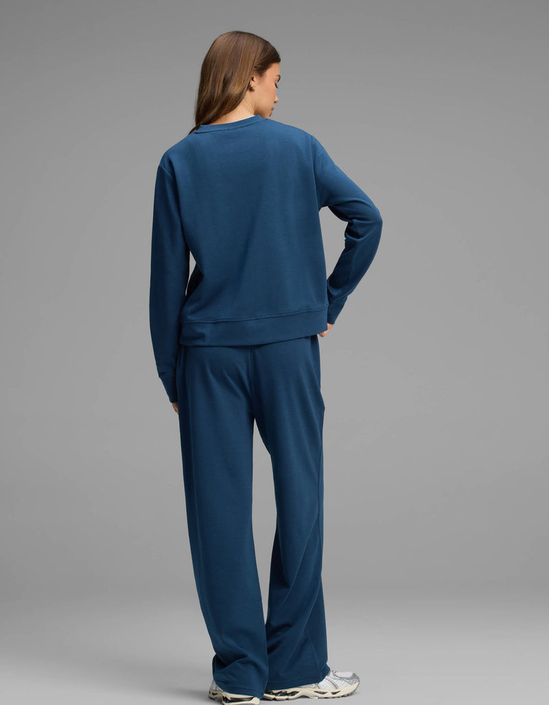 A person stands facing away, wearing the Cozy Earth Women's Versa Crewneck & Straight Leg Pant Set in blue with white sneakers, against a plain gray background.