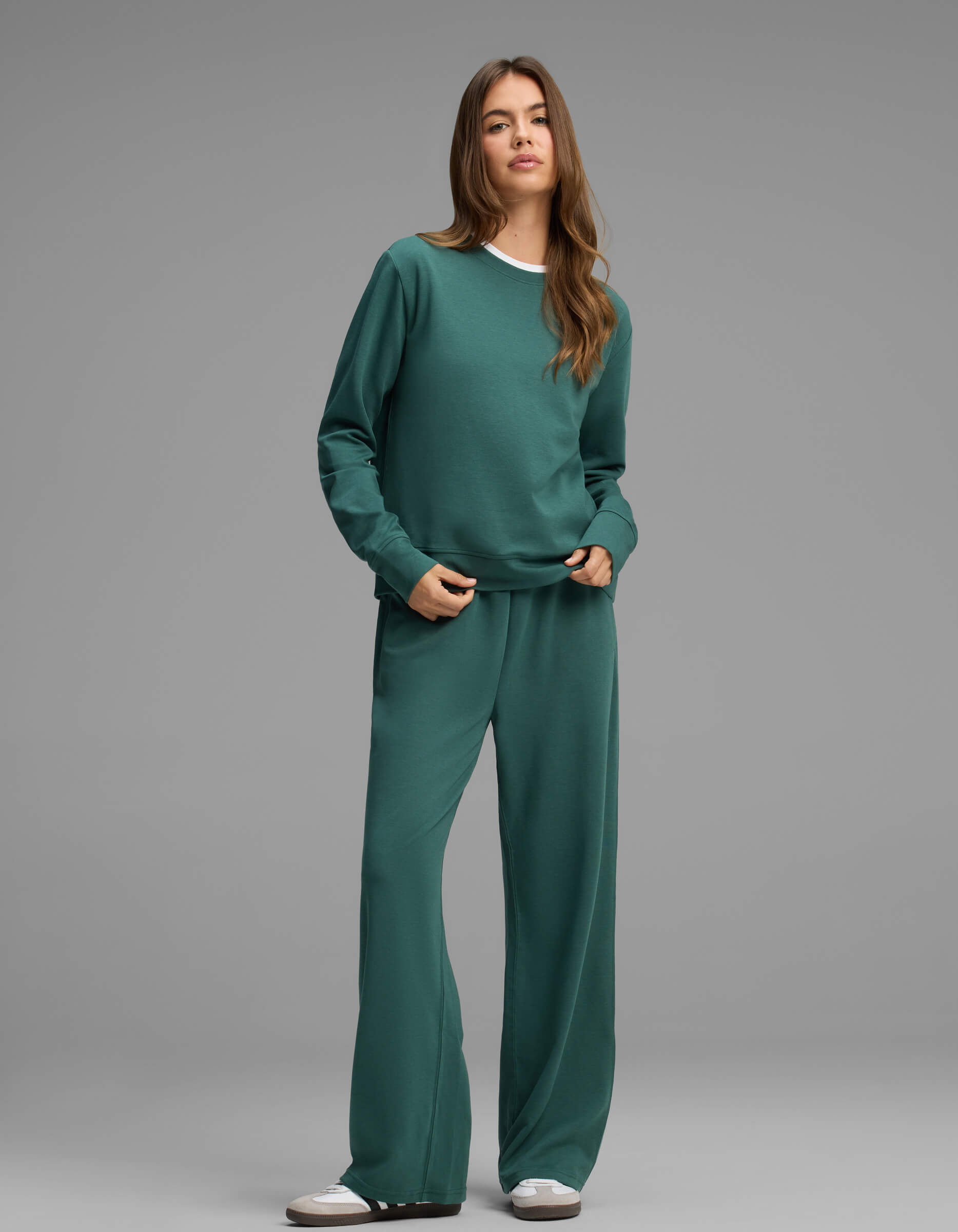 A person stands against a gray background, wearing the Cozy Earth Women's Versa Crewneck & Straight Leg Pant Set in teal, paired with white sneakers.|Color:Uniform