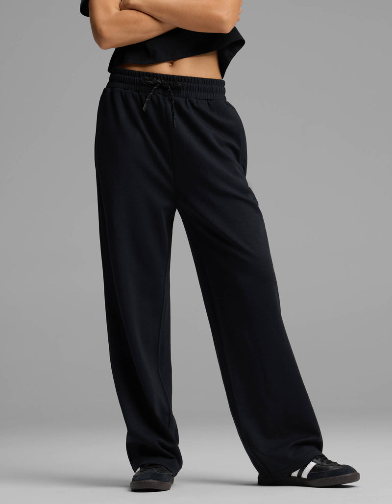 A person stands with arms crossed, dressed in Cozy Earth's Women's Versa Straight Leg Pant, paired with a black top and black sneakers with white accents against a plain gray background. Their face is not visible in the image.