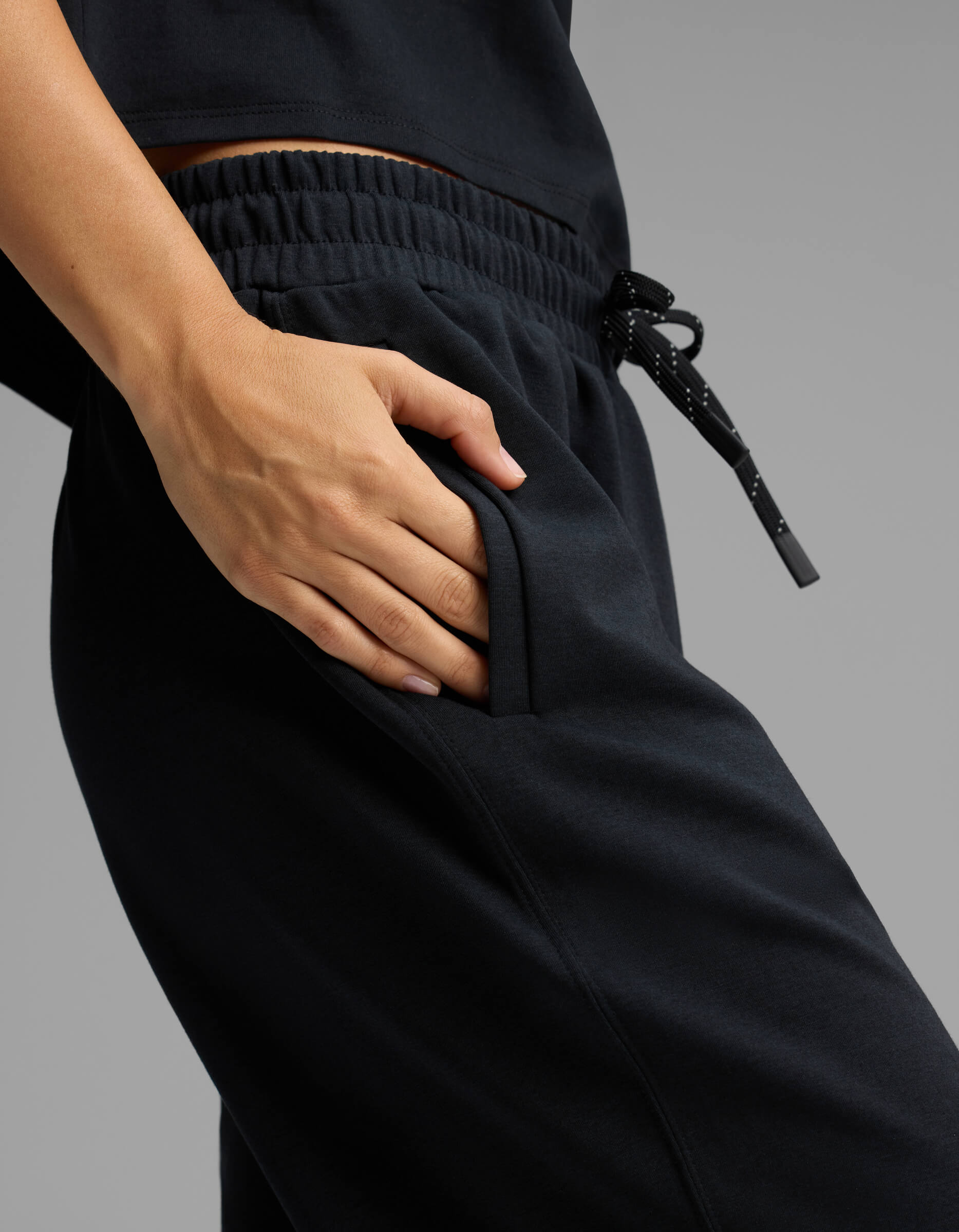 A person wearing Cozy Earth's Women's Versa Straight Leg Pant in black, paired with a matching black top, has their right hand in the pocket of their pants. The pants feature an elastic waistband and a drawstring with white dotted accents.