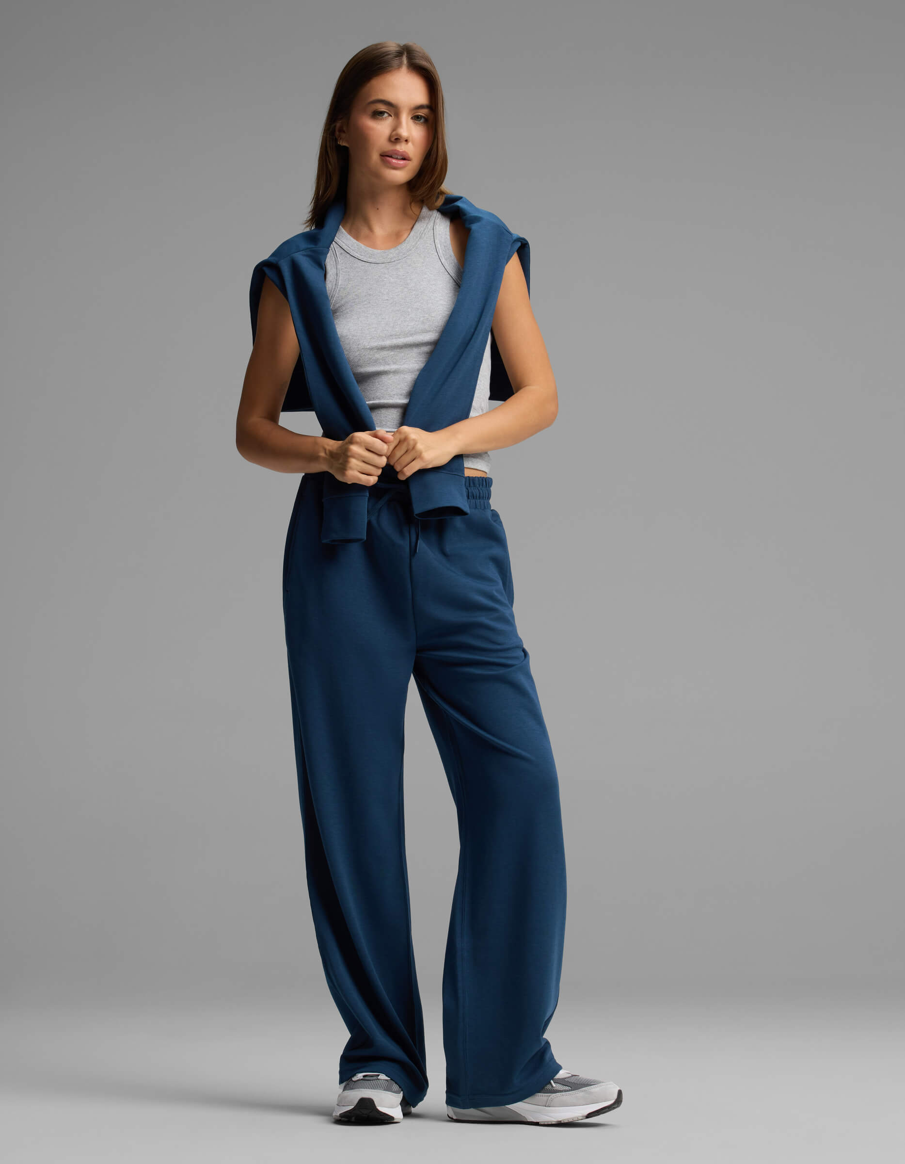 A woman stands against a plain gray background, looking forward with a relaxed posture, wearing Cozy Earth's Women's Versa Straight Leg Pant in blue, paired with a gray tank top and a matching blue sweatshirt draped over her shoulders.
