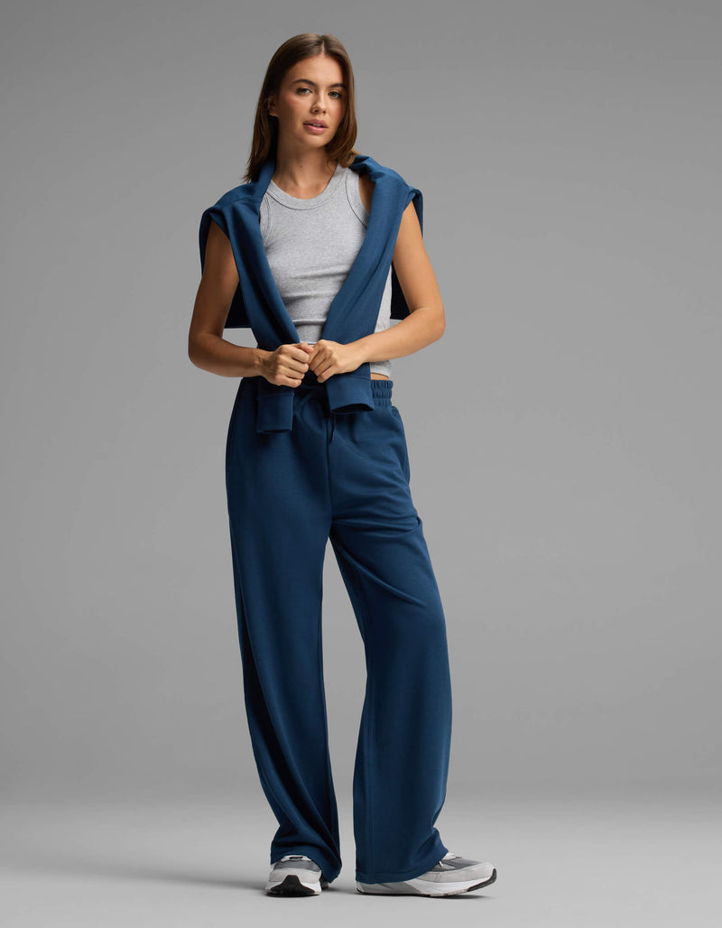 A woman stands against a plain gray background, looking forward with a relaxed posture, wearing Cozy Earth's Women's Versa Straight Leg Pant in blue, paired with a gray tank top and a matching blue sweatshirt draped over her shoulders.